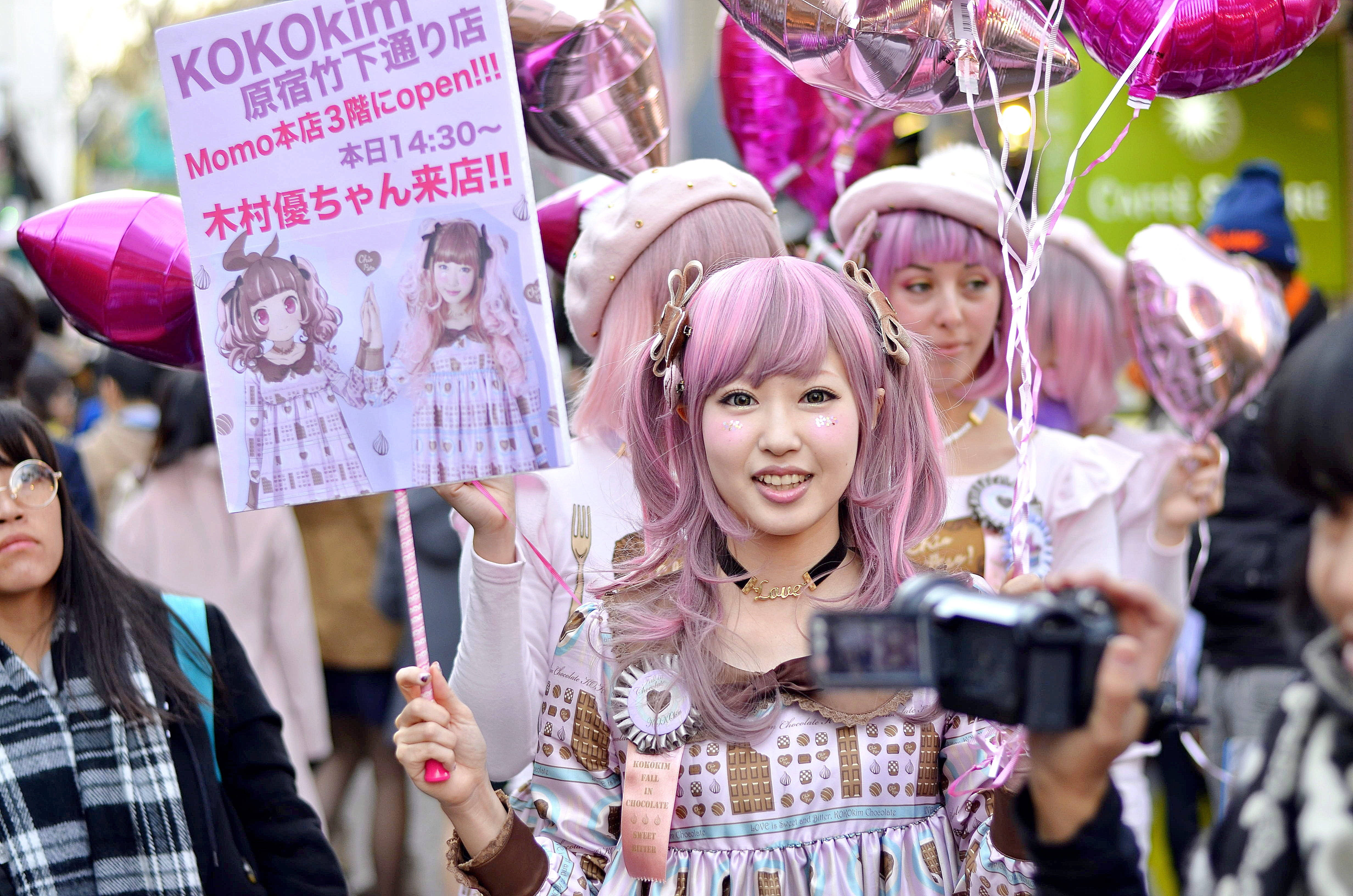 東京 桌布 壁紙 — 一位微笑的年輕女性，粉紅色頭髮，穿著蘿莉塔服裝，在擁擠的街頭活動中拿著標語牌和氣球。