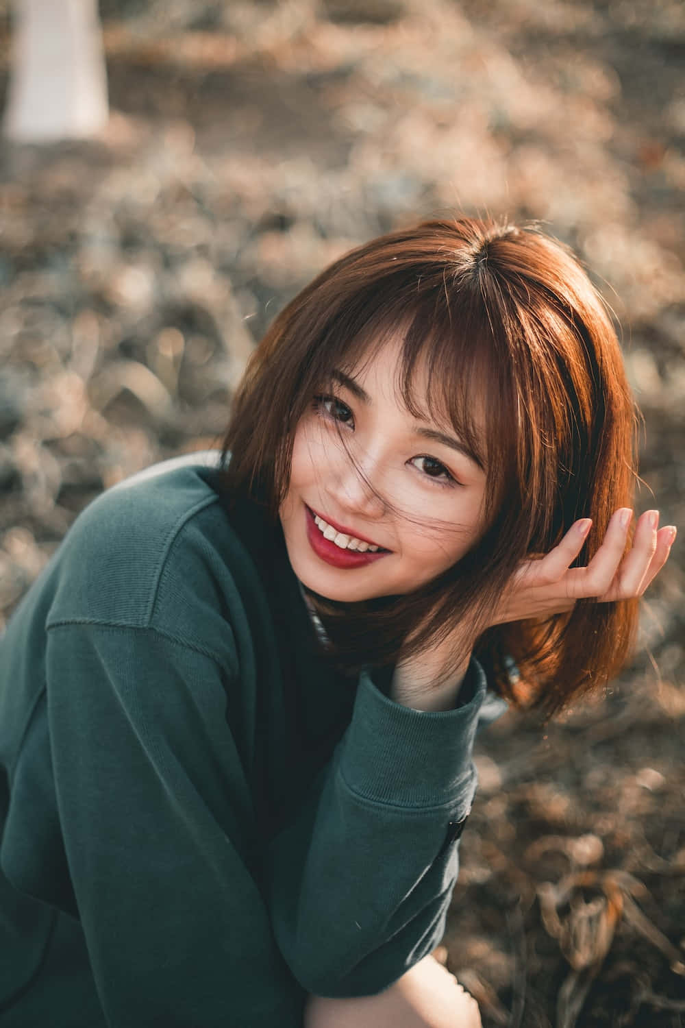 美女 桌布 壁紙 — 一位微笑的年輕女性，紅褐色頭髮和紅色唇膏，穿著綠色運動衫在戶外擺姿勢，背景是模糊的大地色。