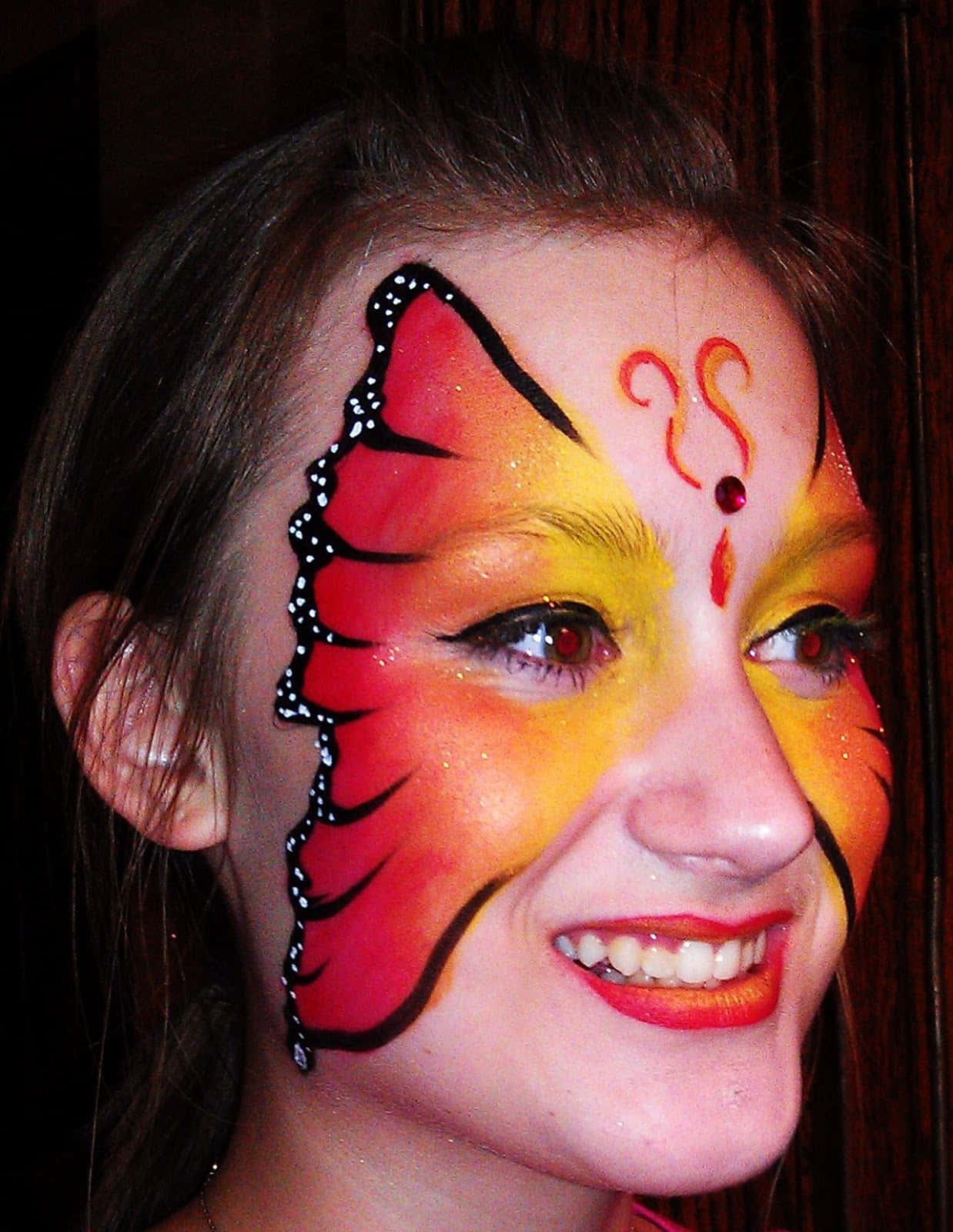 Butterfly Face Painting 桌布 壁紙 — 一位微笑的年輕女性，臉上繪有紅、黃、黑色的鮮豔蝴蝶圖案，背景為深色木質。