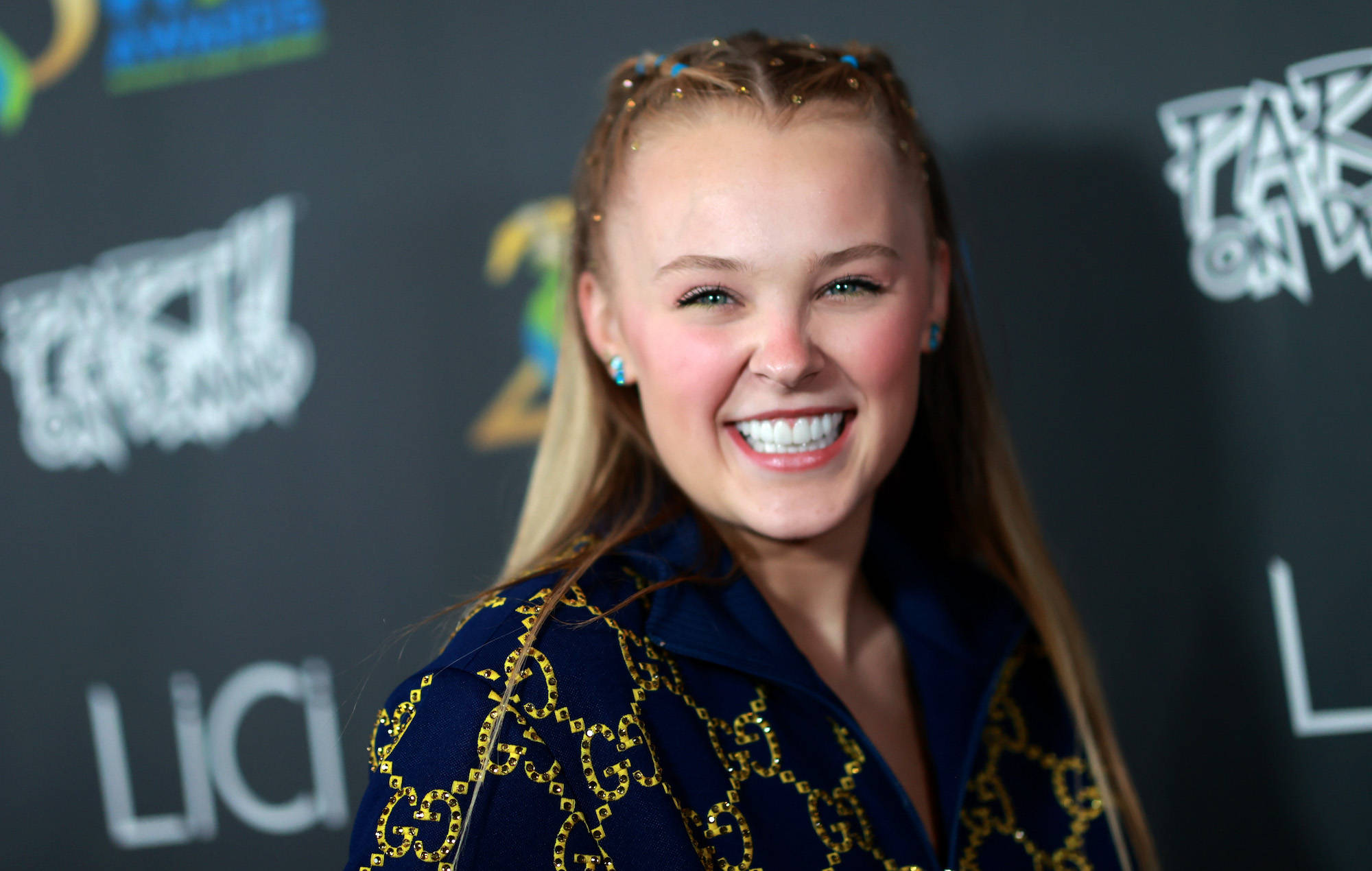 JoJo Siwa 桌布 壁紙 — 一位微笑的年輕女性，金髮藍耳環，身穿海軍藍外套，帶有金色鏈條刺繡，在紅地毯活動中。