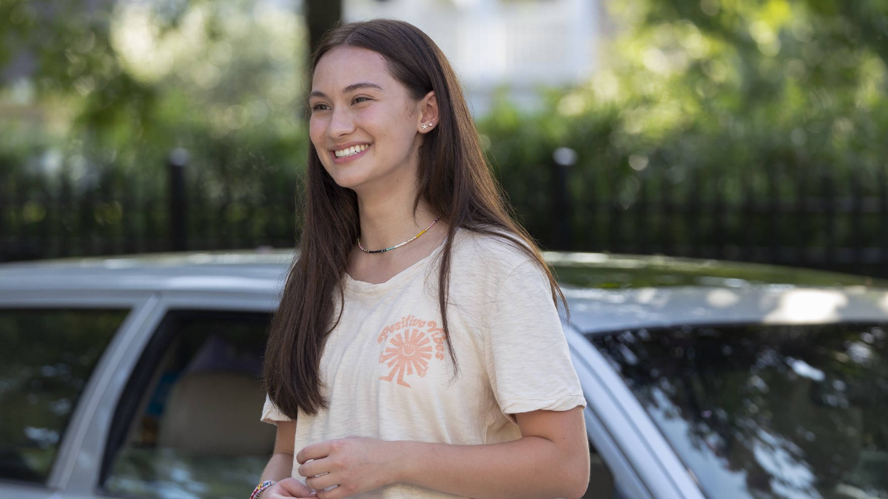 Teenage Girl 桌布 壁紙 — 一位微笑的年輕女性，長長的棕色頭髮，站在一輛銀色車旁邊，在陽光普照的戶外環境中。