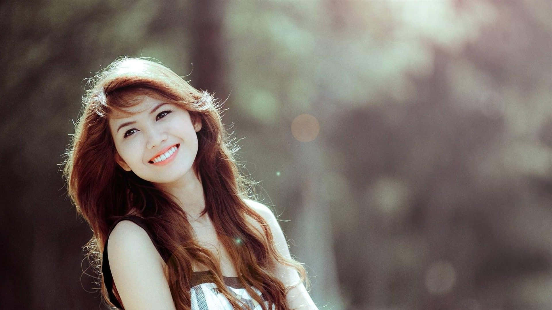 Beautiful Girls 桌布 壁紙 — 一位微笑的年輕女性，長長的棕色頭髮，站在陽光普照的森林中，背景是柔和的散景效果。