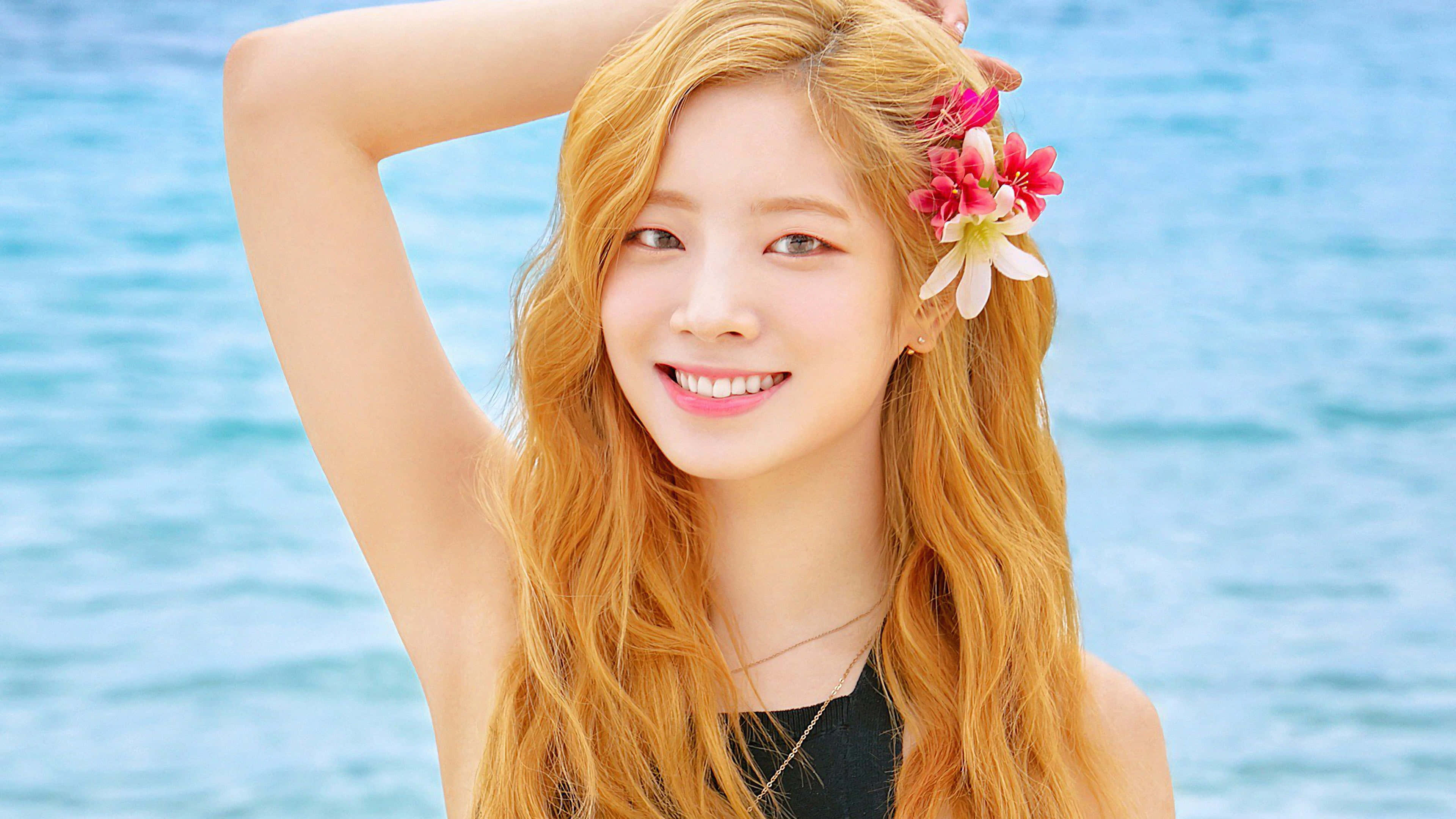 twice 桌布 壁紙 — 一位微笑的年輕女性，長長的金髮上點綴著粉紅色和白色的花朵，站在明亮的藍色海洋背景下。