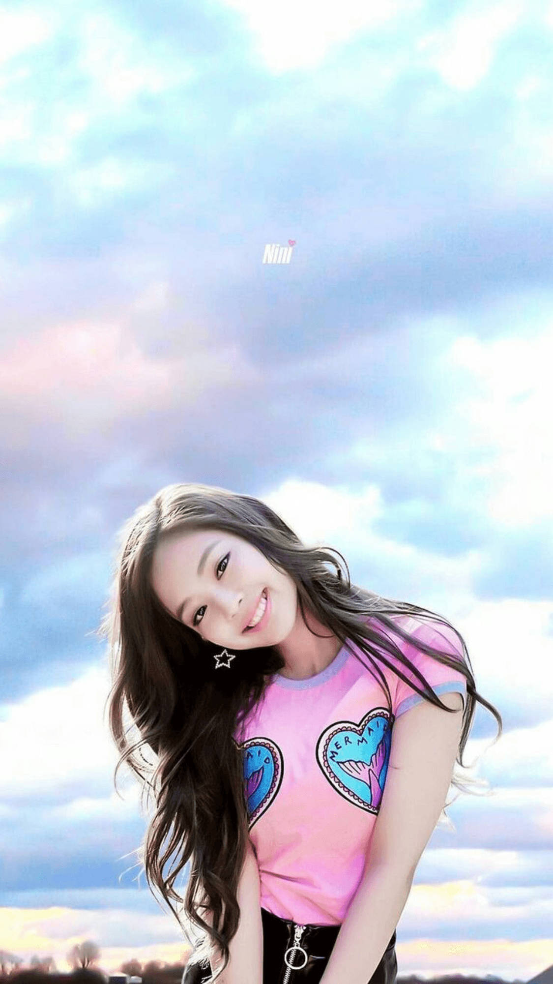 Jennie 桌布 壁紙 — 一位微笑的年輕女性，長長的黑髮，穿著粉紅色心形印花T恤，背景是柔和粉彩的天空，散落著雲朵。