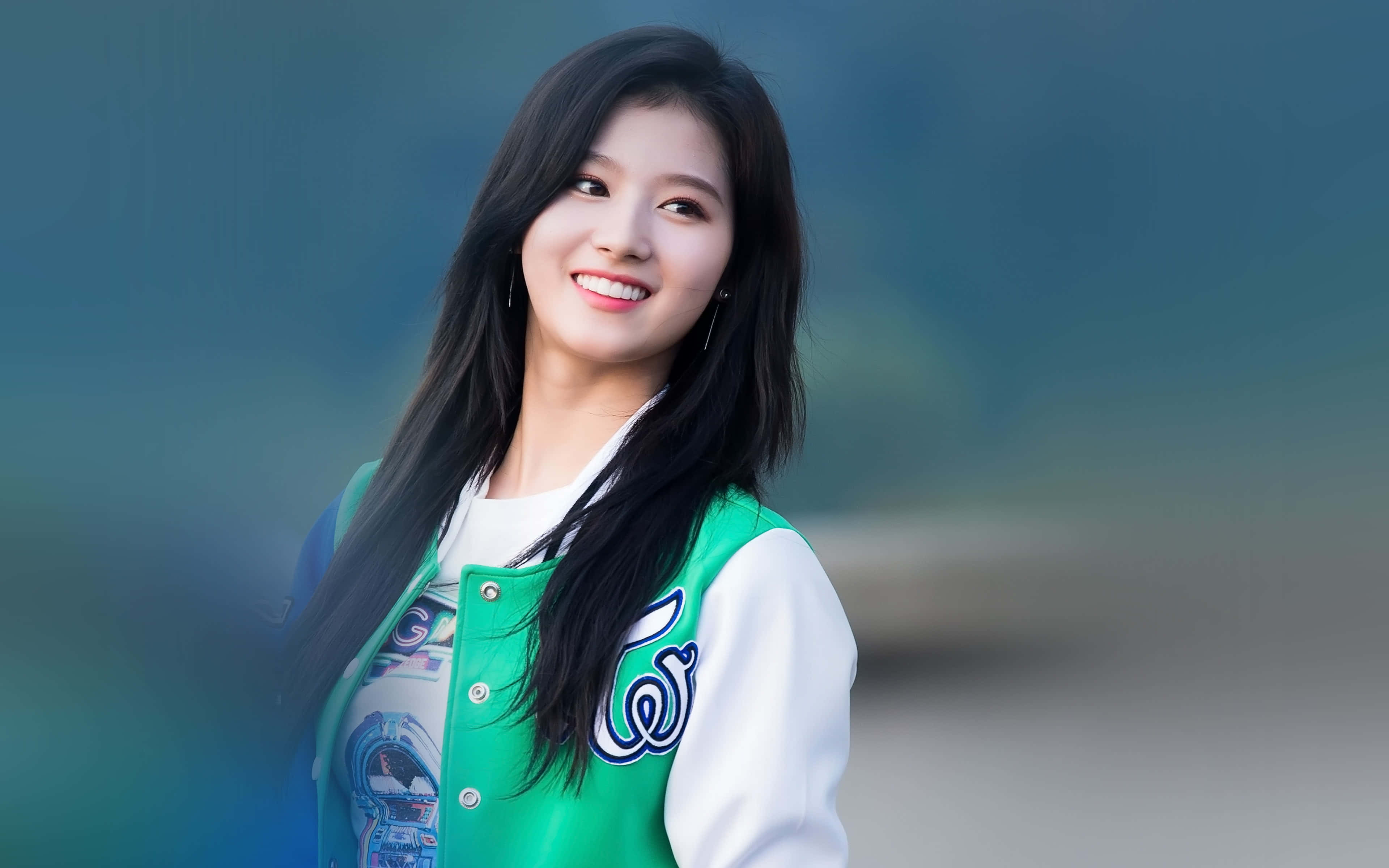 twice 桌布 壁紙 — 一位微笑的年輕女性，長長的黑髮，穿著綠色和白色的校隊夾克，背景是柔和的模糊青綠色。