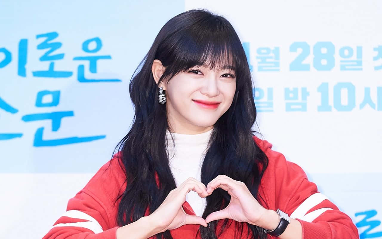Finger Heart 桌布 壁紙 — 一位微笑的年輕女性，長長的黑髮，在淺藍色宣傳背景前，穿著紅色連帽衫，用雙手比出心形。