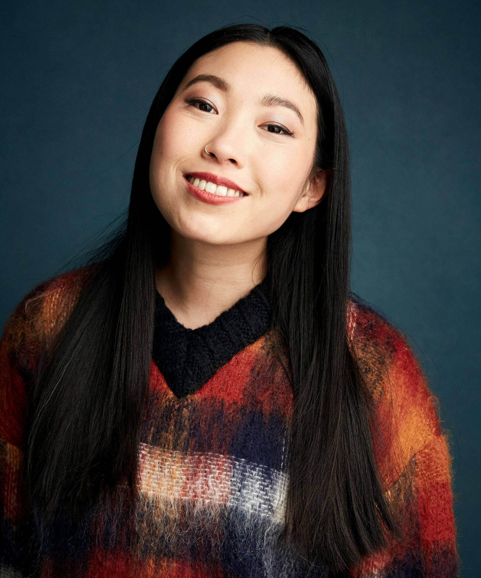 Awkwafina 桌布 壁紙 — 一位微笑的年輕女性，長長的黑髮，穿著色彩繽紛的質感毛衣，背景是深藍色的攝影棚。
