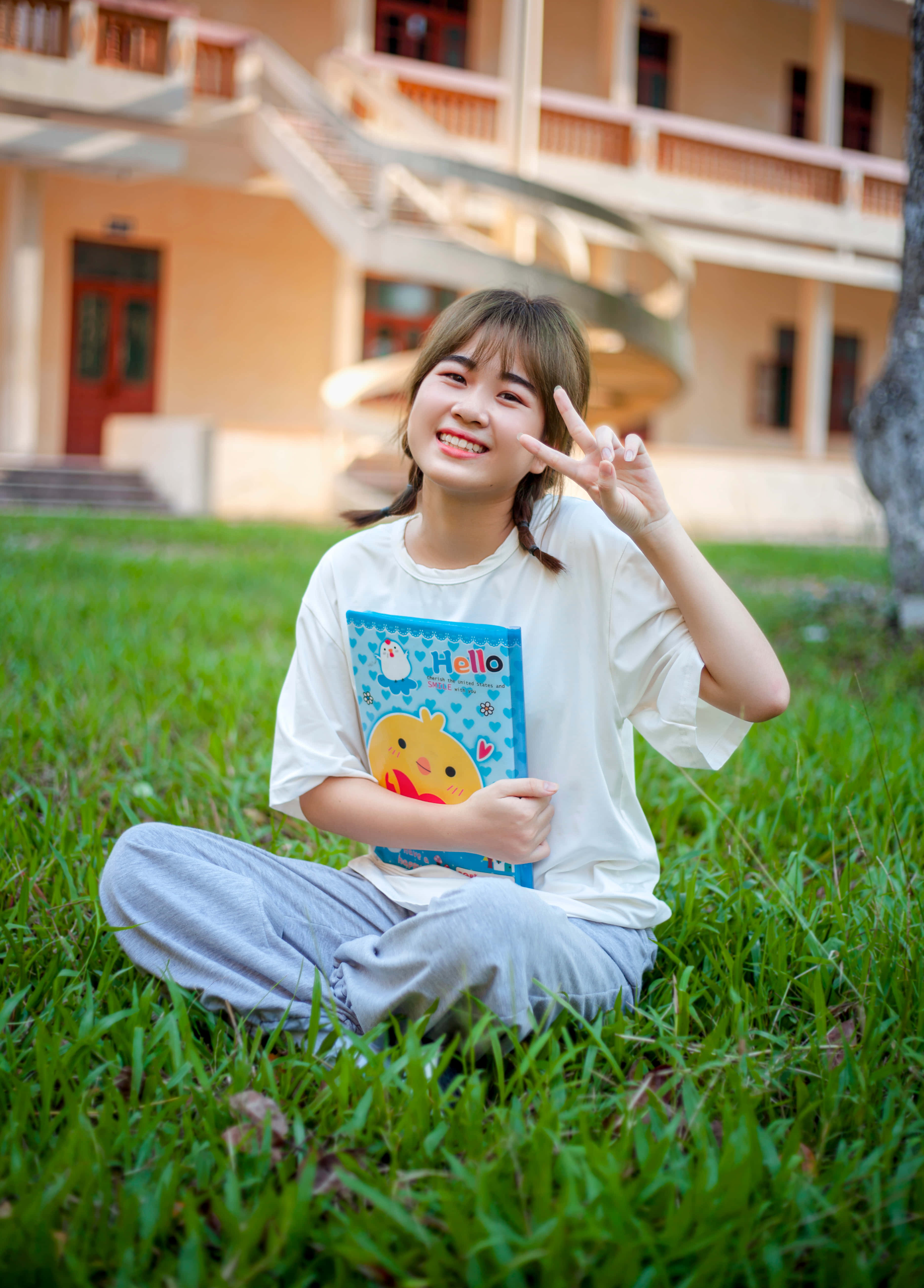 Cute Girl 桌布 壁紙 — 一位微笑的年輕女性盤腿坐在米色建築前充滿活力的綠色草地上，手持彩色筆記本並做出和平手勢。