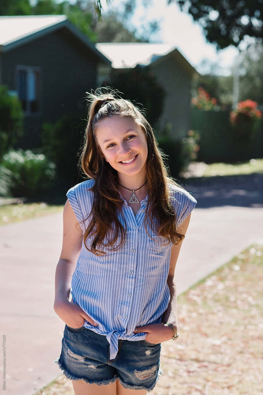 teenager 桌布 壁紙 — 一位微笑的年輕女性穿著條紋襯衫和牛仔短褲，在陽光明媚的日子裡站在戶外，背景是房屋和樹木。