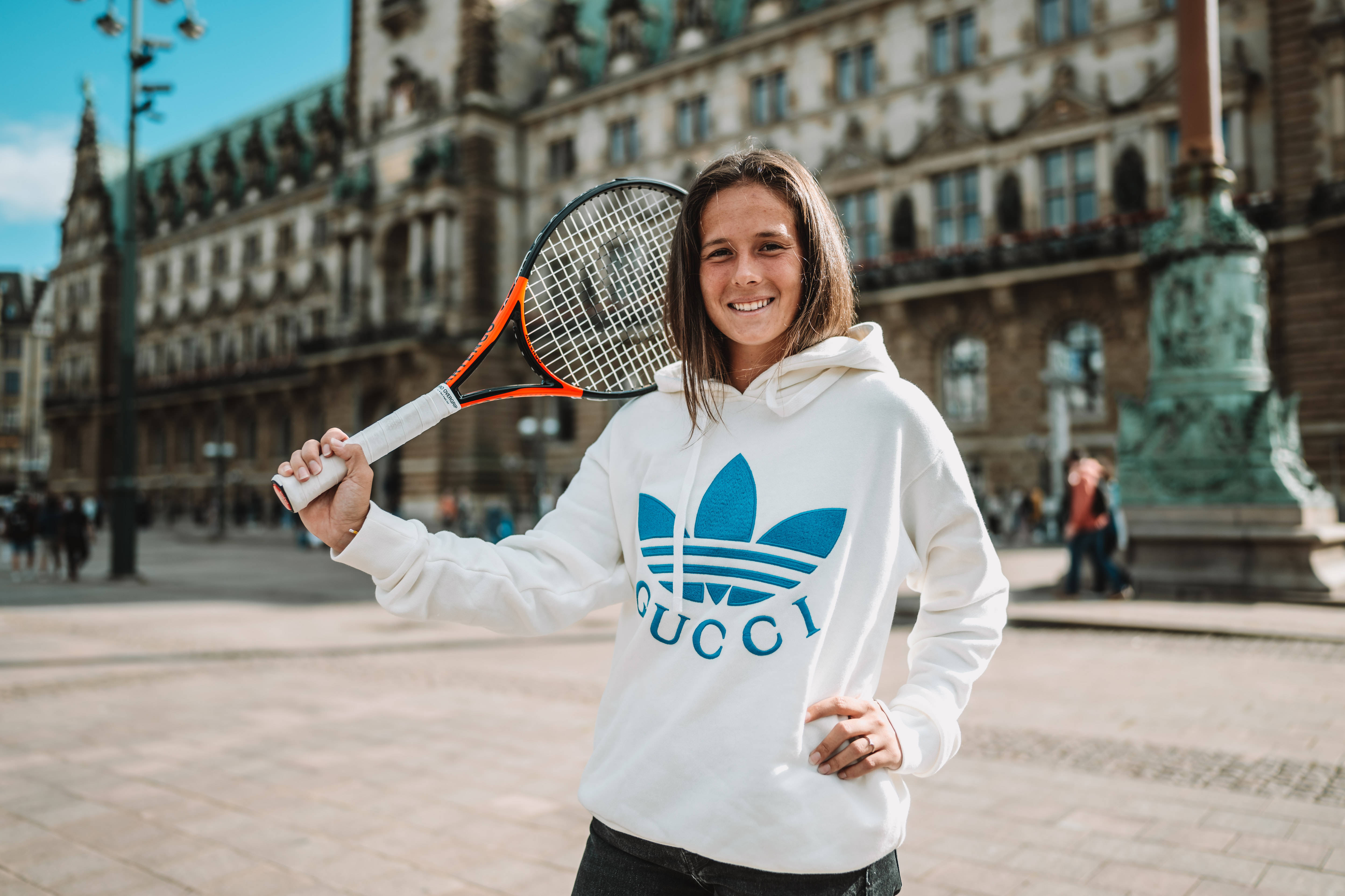 Athletic 桌布 壁紙 — 一位微笑的年輕女性穿著白色Gucci連帽衫，在陽光普照的歐洲廣場上拿著網球拍，周圍是歷史建築。