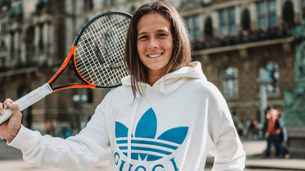 Athletic 桌布 壁紙 — 一位微笑的年輕女性穿著白色連帽衫，在歷史建築背景下手持網球拍。