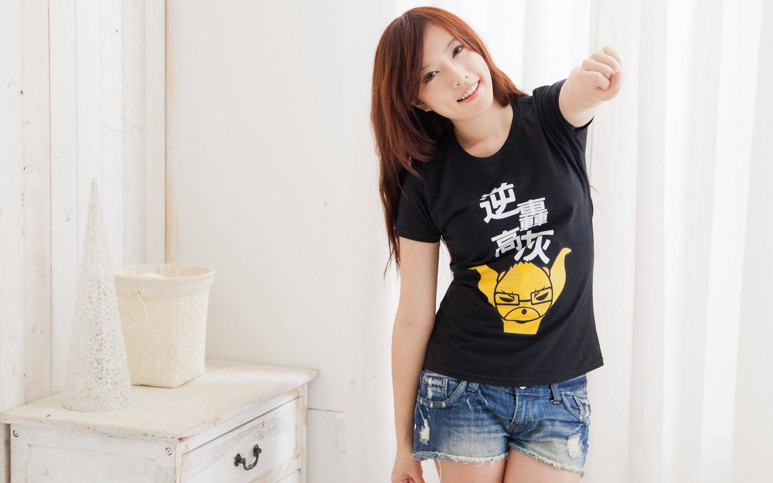 女孩 桌布 壁紙 — 一位微笑的年輕女性穿著黑色T恤，上面有黃色卡通圖案和白色文字，在室內靠近白色抽屜櫃和窗簾處俏皮地擺姿勢。
