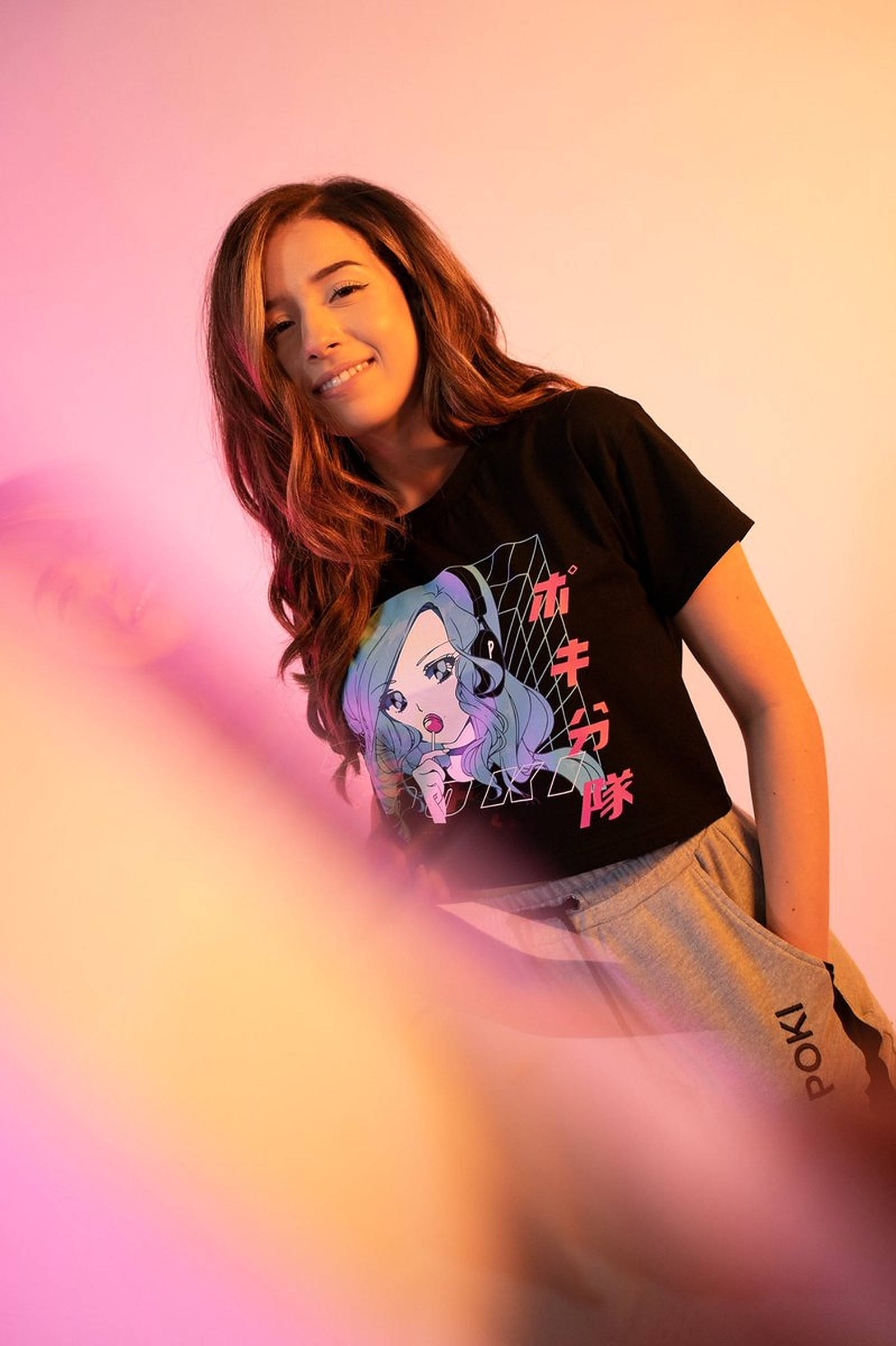 Pokimane 桌布 壁紙 — 一位微笑的年輕女性穿著黑色動漫圖案T恤，在柔和的粉紅和橙色漸變背景前擺姿勢，前景元素模糊。