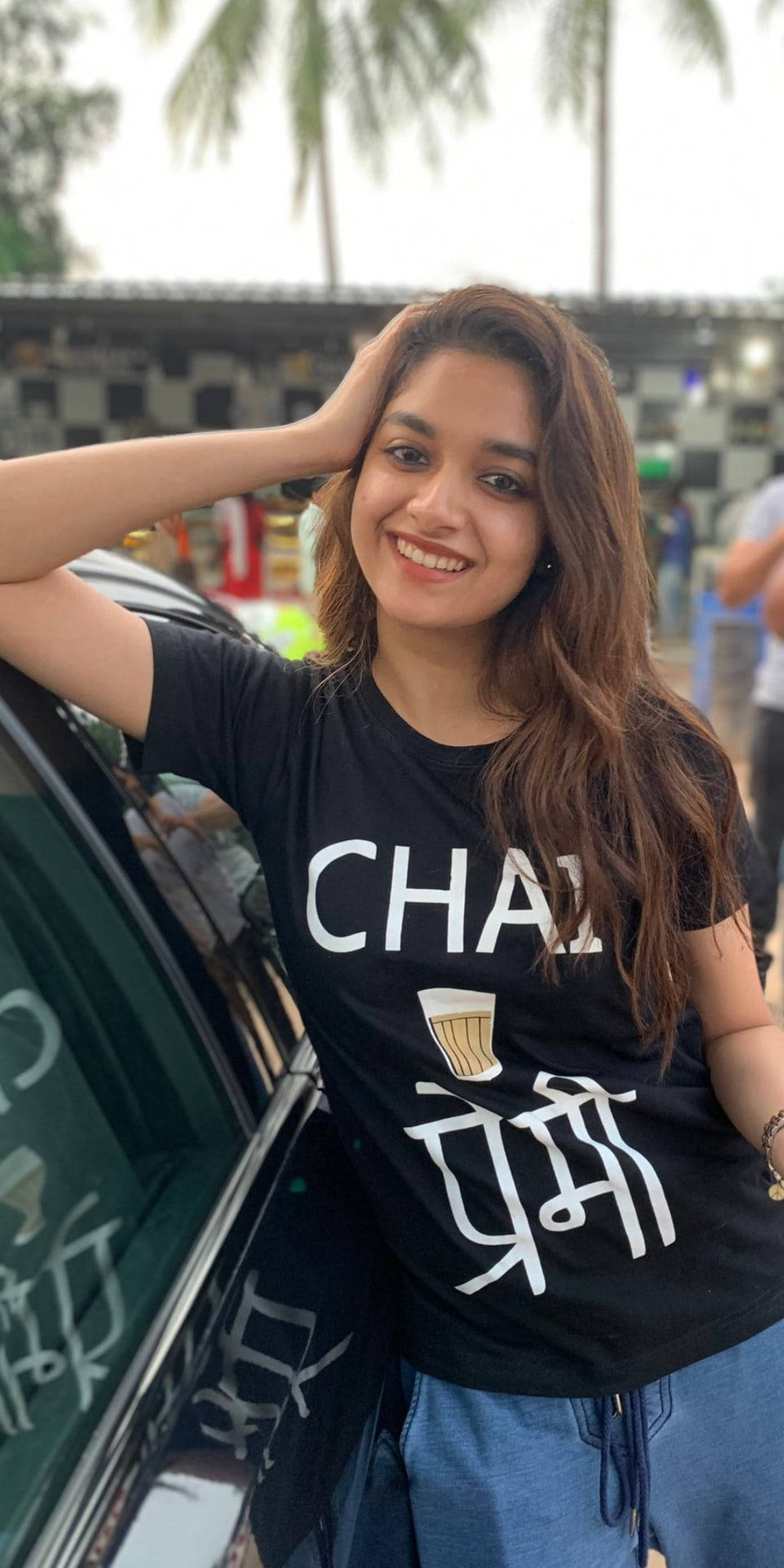 telugu 桌布 壁紙 — 一位微笑的年輕女性穿著黑色奶茶主題T恤，隨意地靠在一輛車上，背景有棕櫚樹。