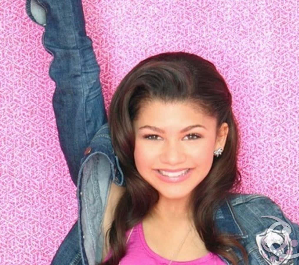 Zendaya 桌布 壁紙 — 一位微笑的年輕女性身穿牛仔外套和粉紅色上衣，在充滿活力的粉紅色圖案背景前舉起一隻手臂擺姿勢。