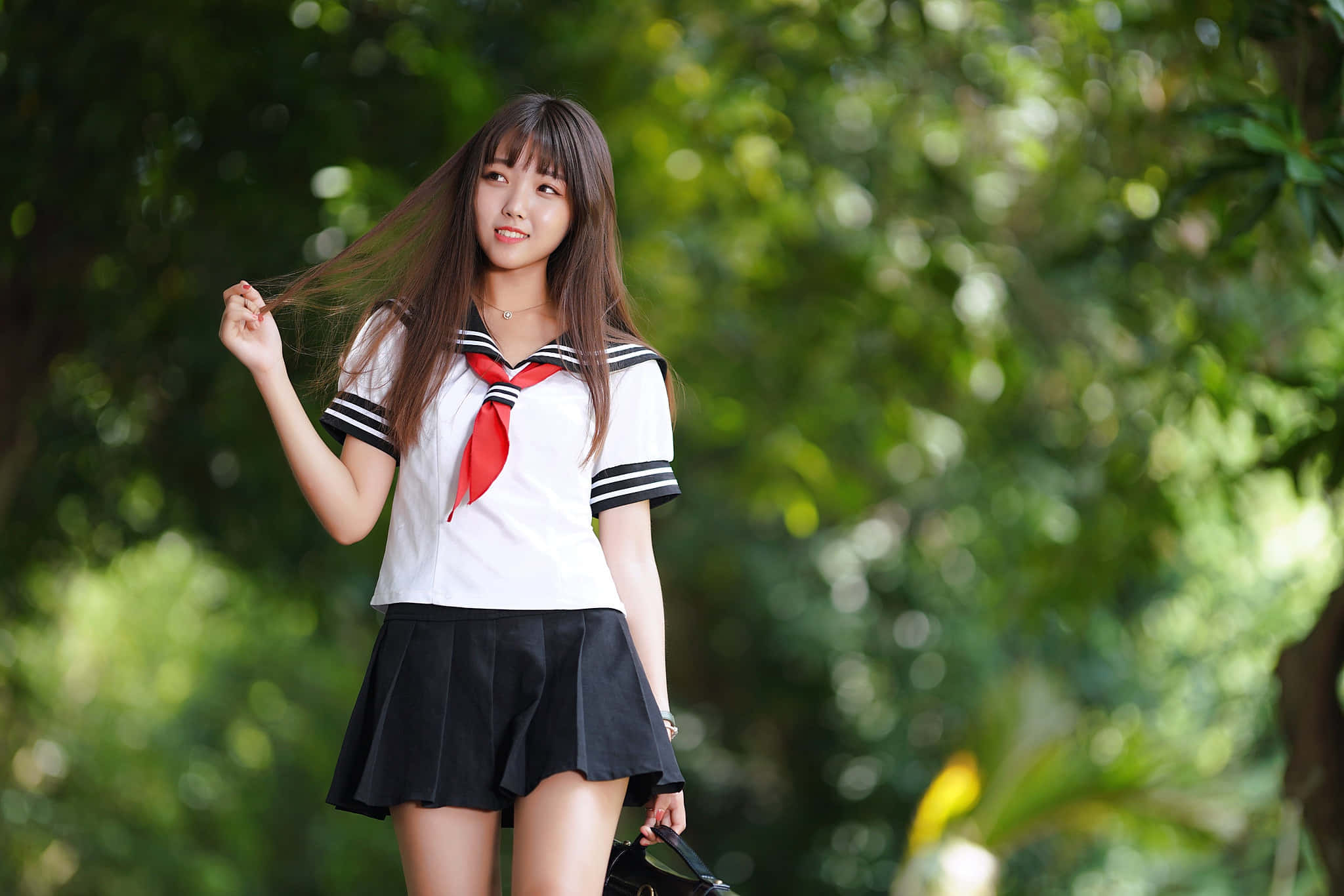 Japanese School Girl 桌布 壁紙 — 一位微笑的年輕女性身穿白色水手服配紅色領帶，站在茂密的綠葉中，輕輕撩起她的長棕髮。