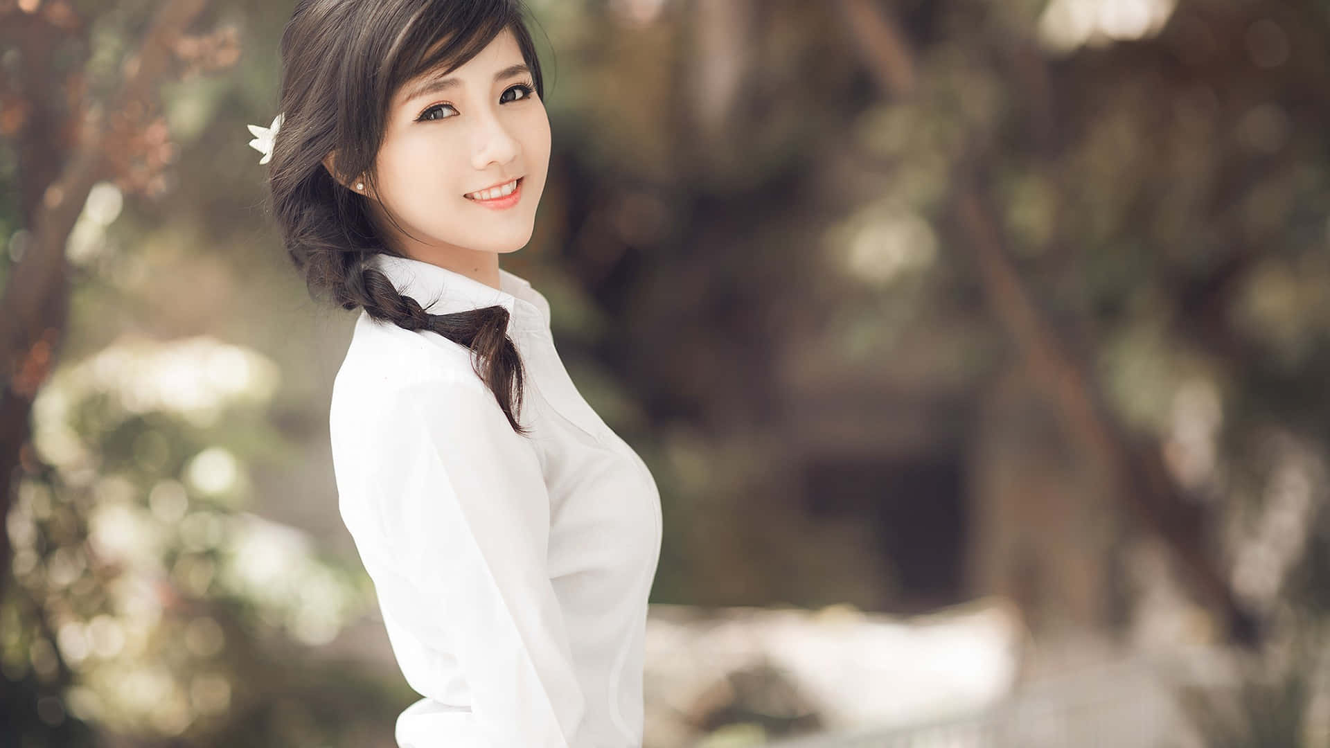 美女 桌布 壁紙 — 一位微笑的年輕女性身穿白色襯衫，站在戶外，柔和的陽光透過她身後模糊的綠色樹葉灑落下來。