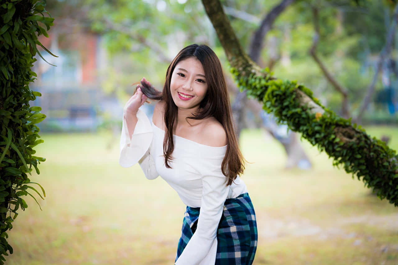 smiling 桌布 壁紙 — 一位微笑的年輕女性身穿白色露肩上衣和格子裙，在陽光明媚的公園裡，在長滿苔蘚的樹枝下俏皮地擺姿勢。