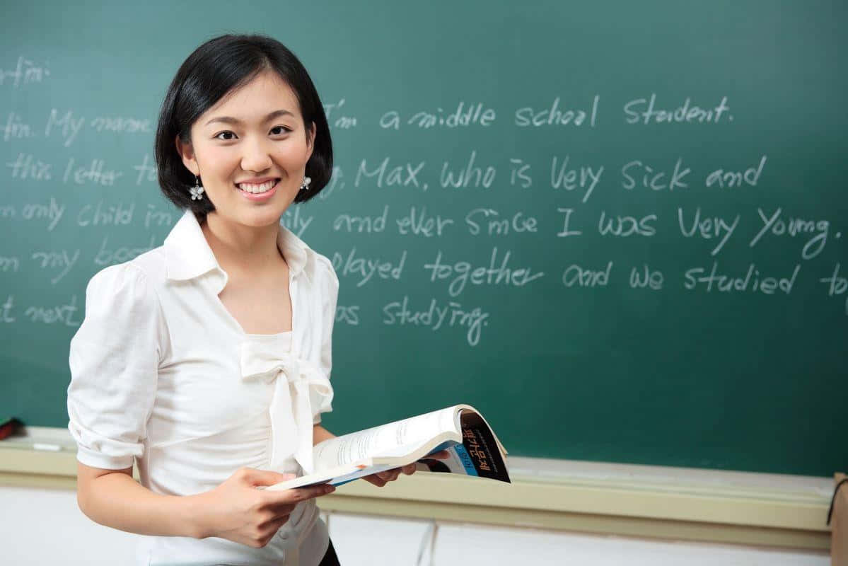 teachers 桌布 壁紙 — 一位微笑的年輕女教師穿著白色襯衫，在綠色黑板前拿著一本書，黑板上有手寫文字。