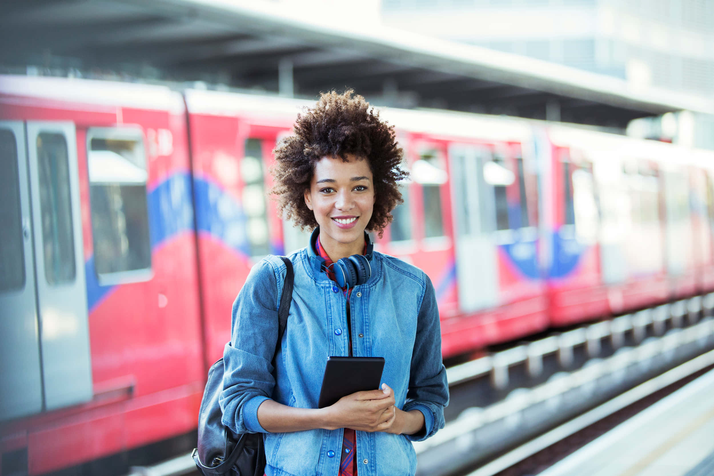 Travel Desktop 桌布 壁紙 — 一位微笑的年輕捲髮女子站在火車站，手持平板電腦和耳機，背景有紅色火車。
