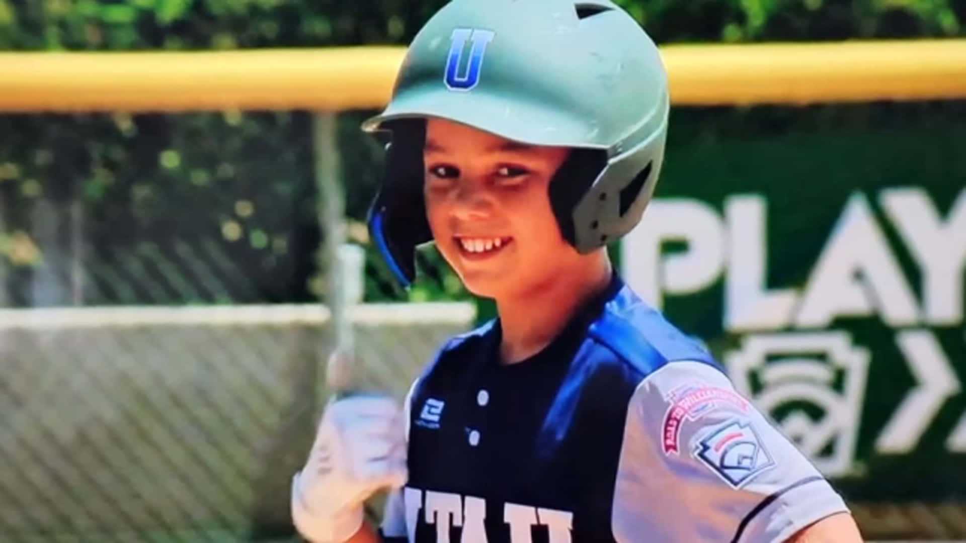 Little League Baseball 桌布 壁紙 — 一位微笑的年輕棒球運動員，身穿藍色制服和頭盔，在明亮的日光下拿著球棒準備比賽。