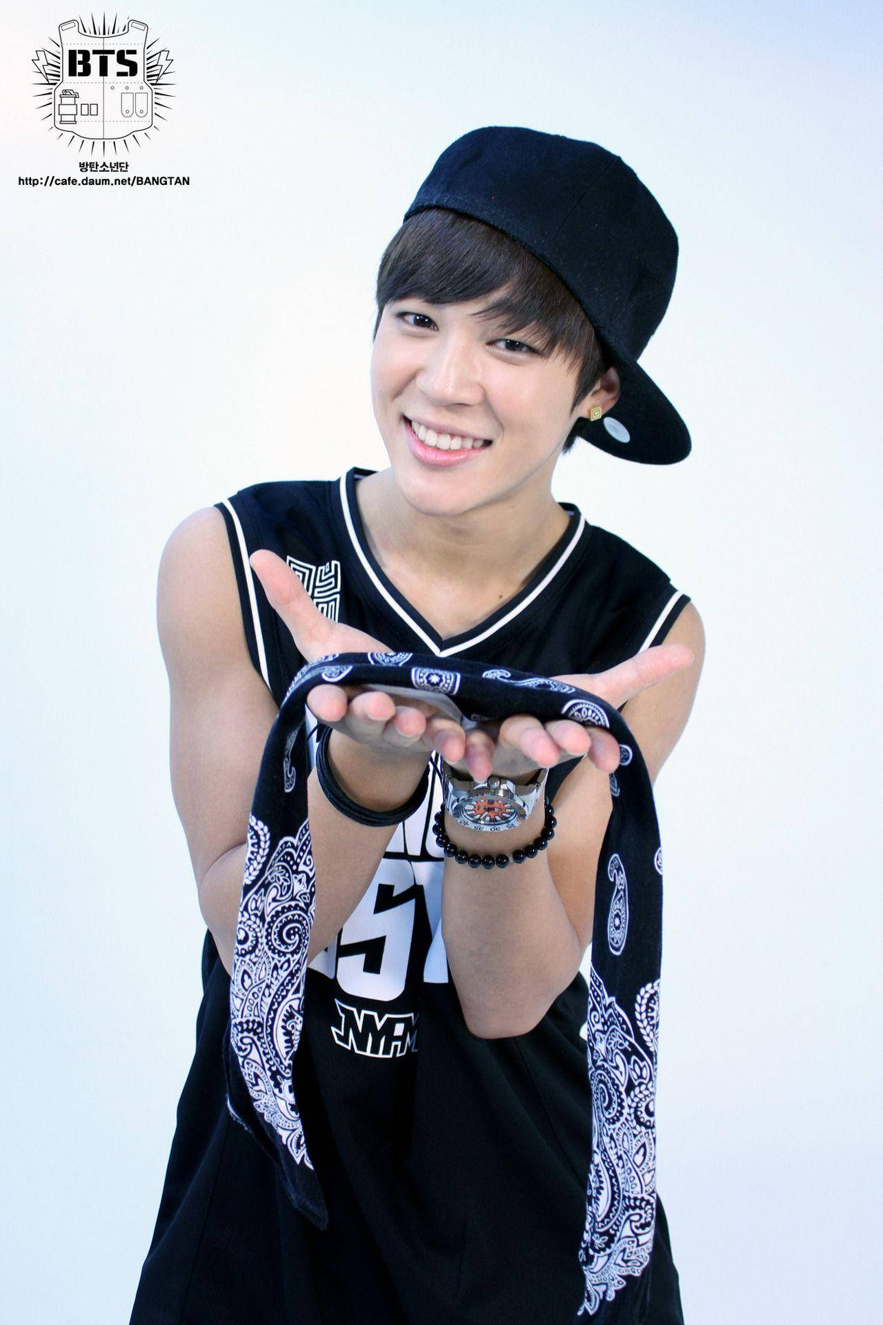 Jimin Bts Cute 桌布 壁紙 — 一位微笑的年輕男子，戴著黑色鴨舌帽和無袖運動衫，在純白色背景前，張開手掌，拿著一條圖案頭巾。