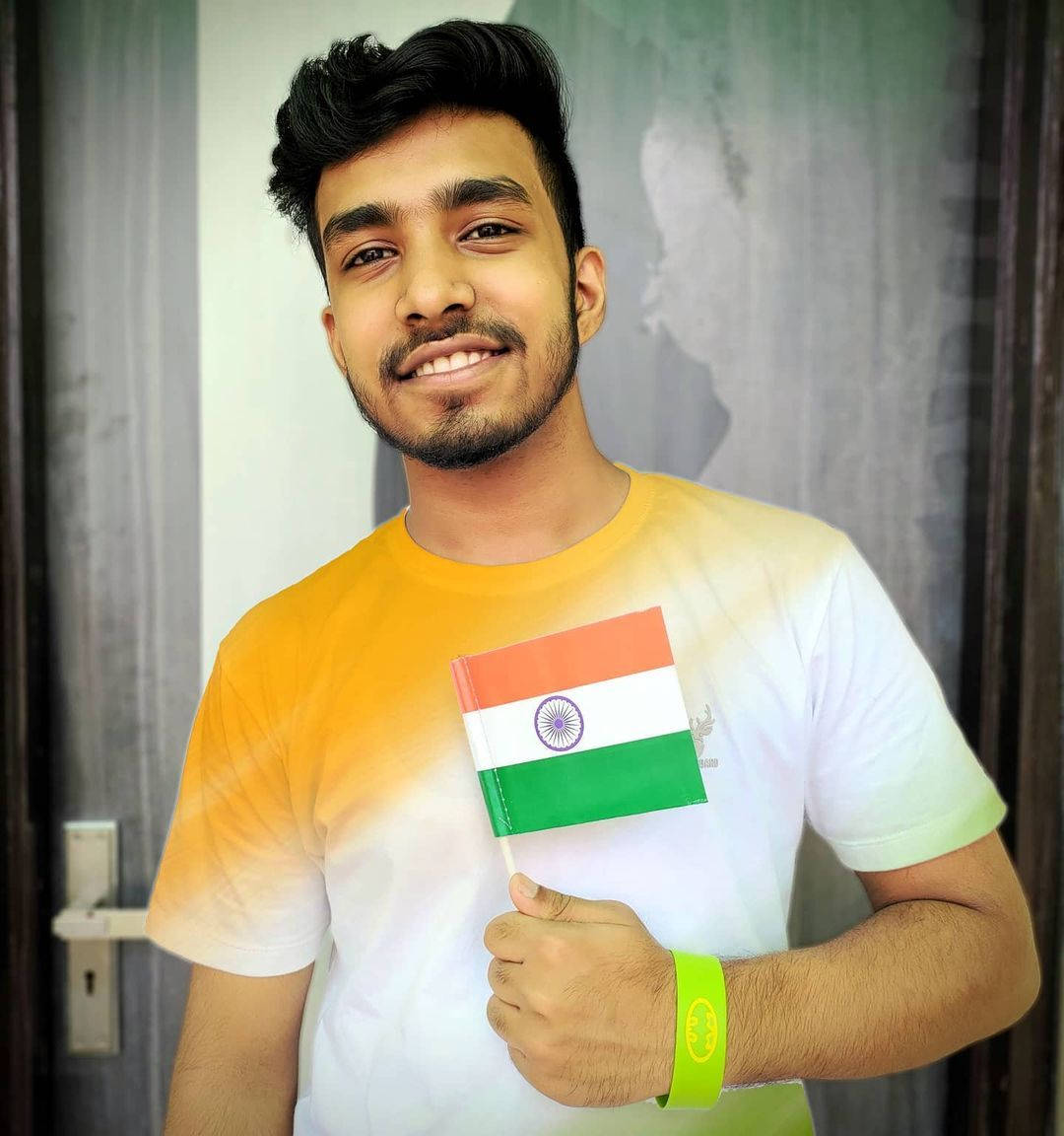 Indian 桌布 壁紙 — 一位微笑的年輕男子，深色頭髮和鬍鬚，穿著漸層黃白綠T恤，手持印度國旗，戴著亮綠色腕帶。