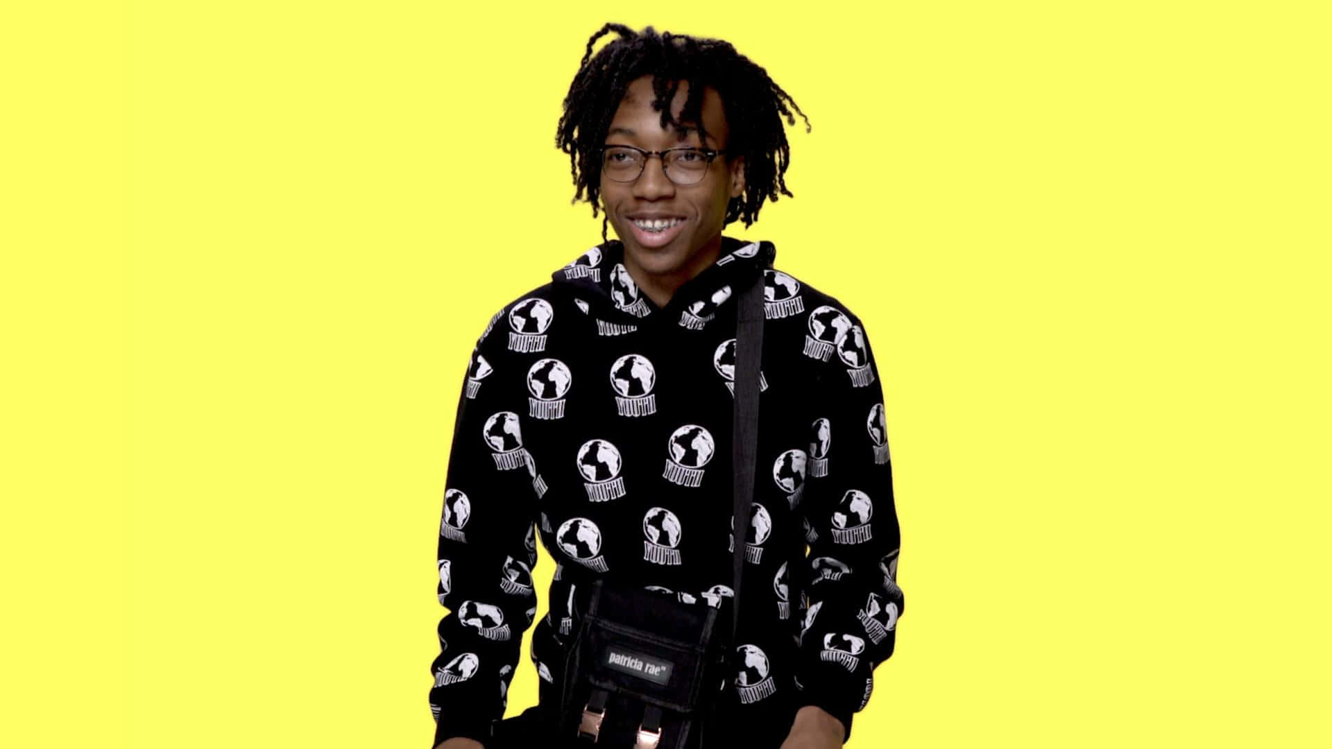 Rappers 桌布 壁紙 — 一位微笑的年輕男子，留著辮子頭，戴著眼鏡，穿著黑色連帽衫，上面有白色地球圖案，背景是明亮的黃色。