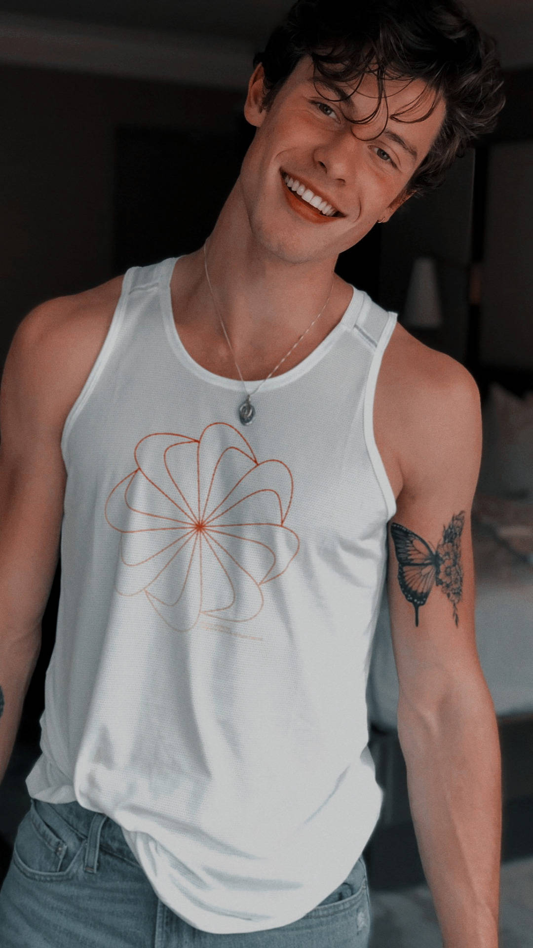 Shawn 桌布 壁紙 — 一位微笑的年輕男子，身穿帶有橙色花卉圖案的白色背心，手臂上有蝴蝶刺青。