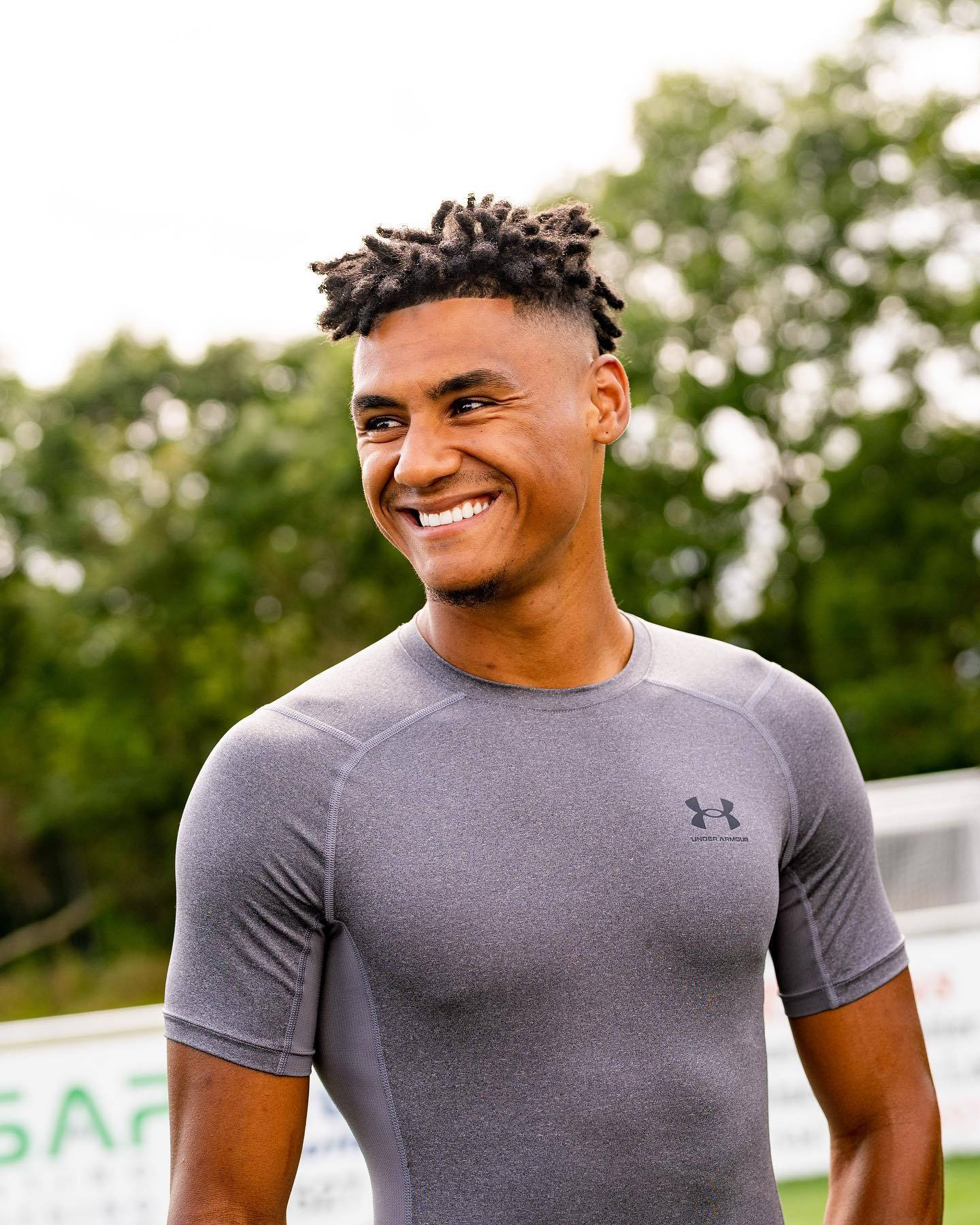 Football Players 桌布 壁紙 — 一位微笑的年輕男子，頭髮有紋理，穿著灰色運動衫，背景是模糊的綠色戶外環境。