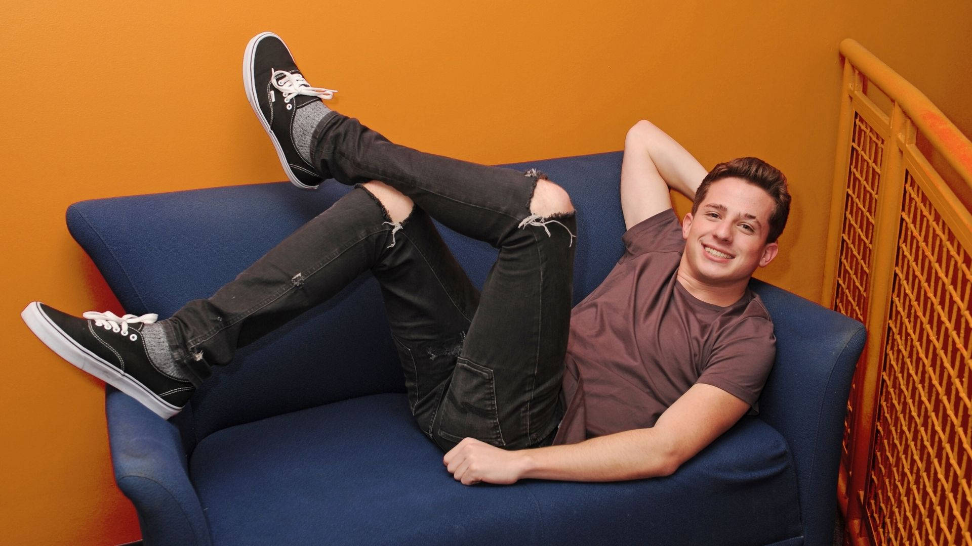 Charlie Puth 桌布 壁紙 — 一位微笑的年輕男子休閒地躺在藍色沙發上，背景是充滿活力的橙色牆壁，穿著破洞牛仔褲和黑色運動鞋。