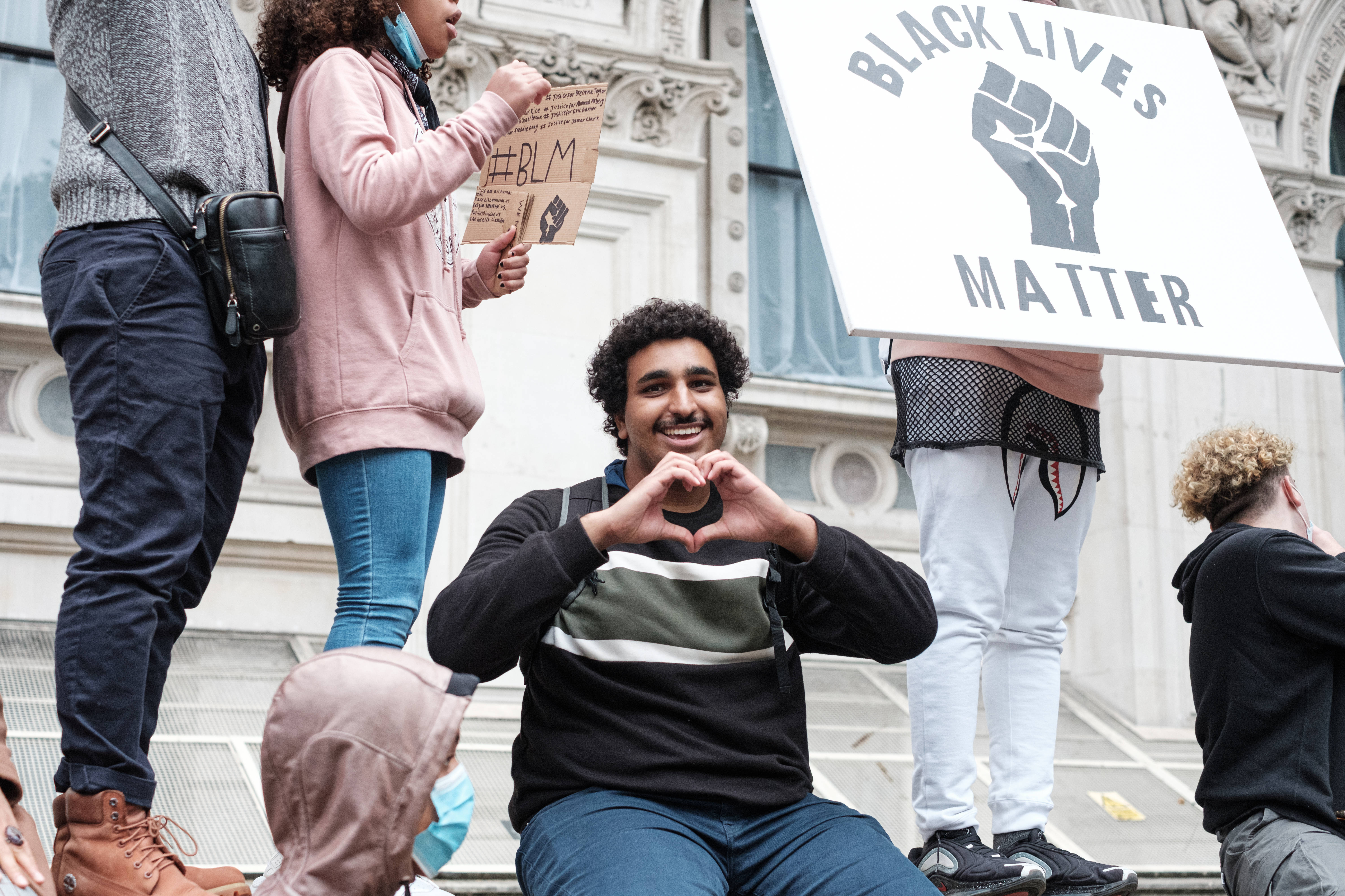 黑色愛情 桌布 壁紙 — 一位微笑的年輕男子在Black Lives Matter抗議活動中坐著，用雙手比出心形，其他人則舉著標語牌。