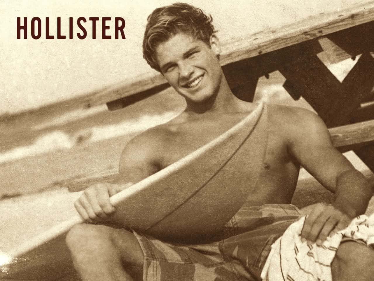 hollister 桌布 壁紙 — 一位微笑的年輕男子穿著圖案泳褲，坐在海灘的木製碼頭上，手持衝浪板。