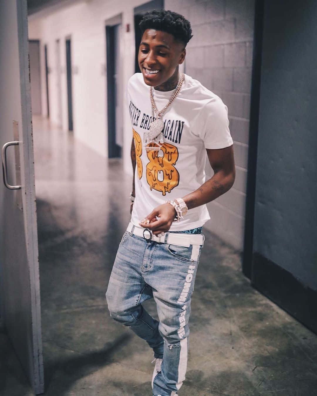 NBA YoungBoy 桌布 壁紙 — 一位微笑的年輕男子穿著白色圖案T恤和牛仔褲，自信地走過煙霧瀰漫的走廊。