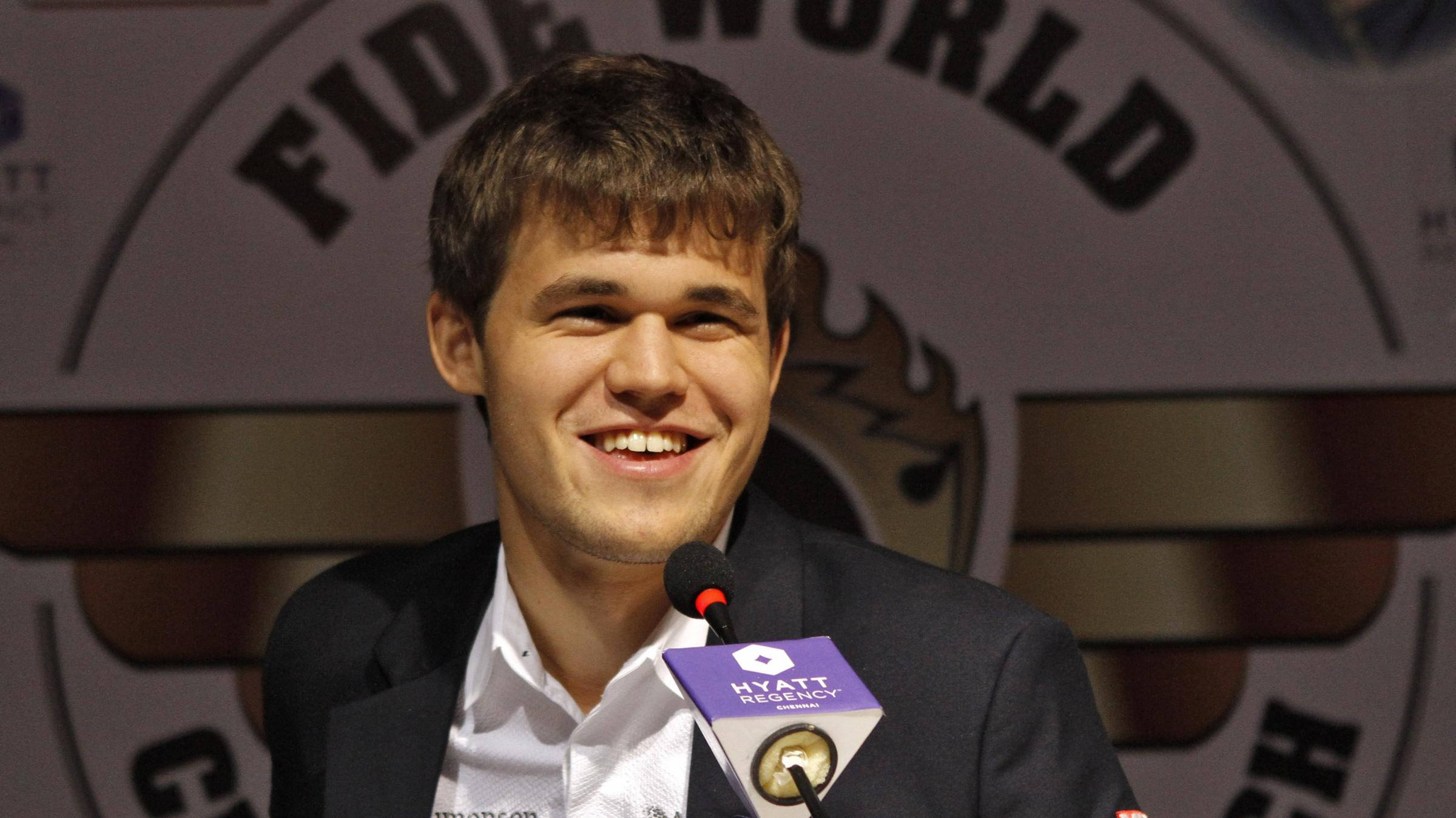 Magnus 桌布 壁紙 — 一位微笑的年輕男子穿著西裝，在FIDE World Chess活動記者會上對著麥克風講話。