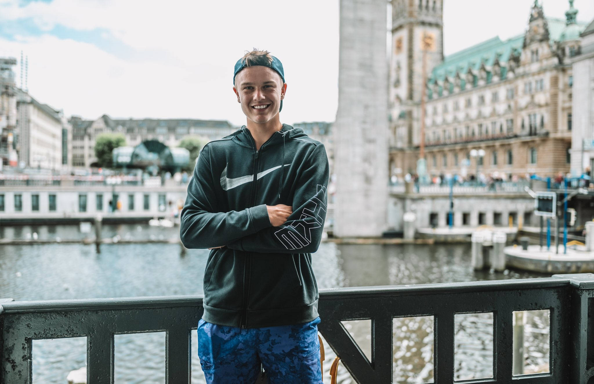 Tennis 桌布 壁紙 — 一位微笑的年輕男子穿著連帽衫和帽子，自信地站在一座橋上，俯瞰著河流，背景是歷史建築。
