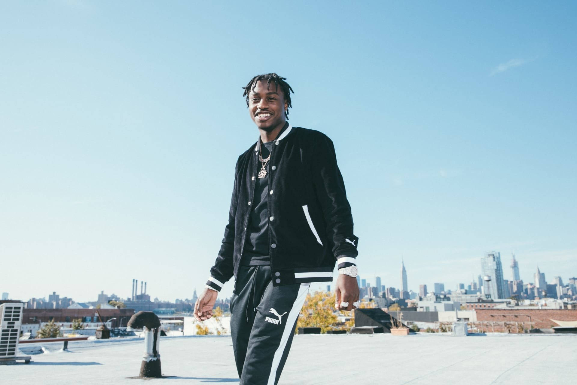 Lil Tjay 桌布 壁紙 — 一位微笑的年輕男子穿著黑色Puma運動服，自信地站在屋頂上，背後是晴朗的藍天和遠處的城市天際線。