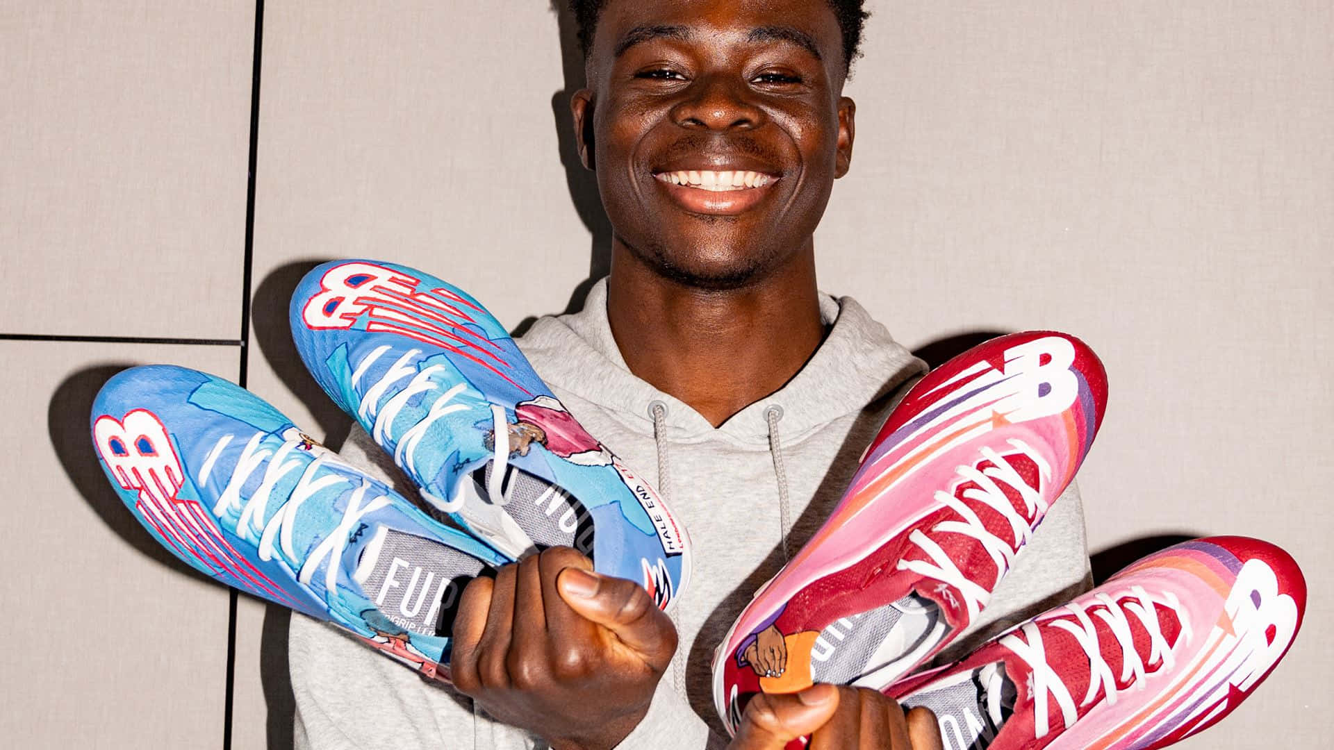 New Balance 桌布 壁紙 — 一位微笑的年輕男子自豪地展示兩雙色彩繽紛的New Balance足球鞋，背景是素牆。