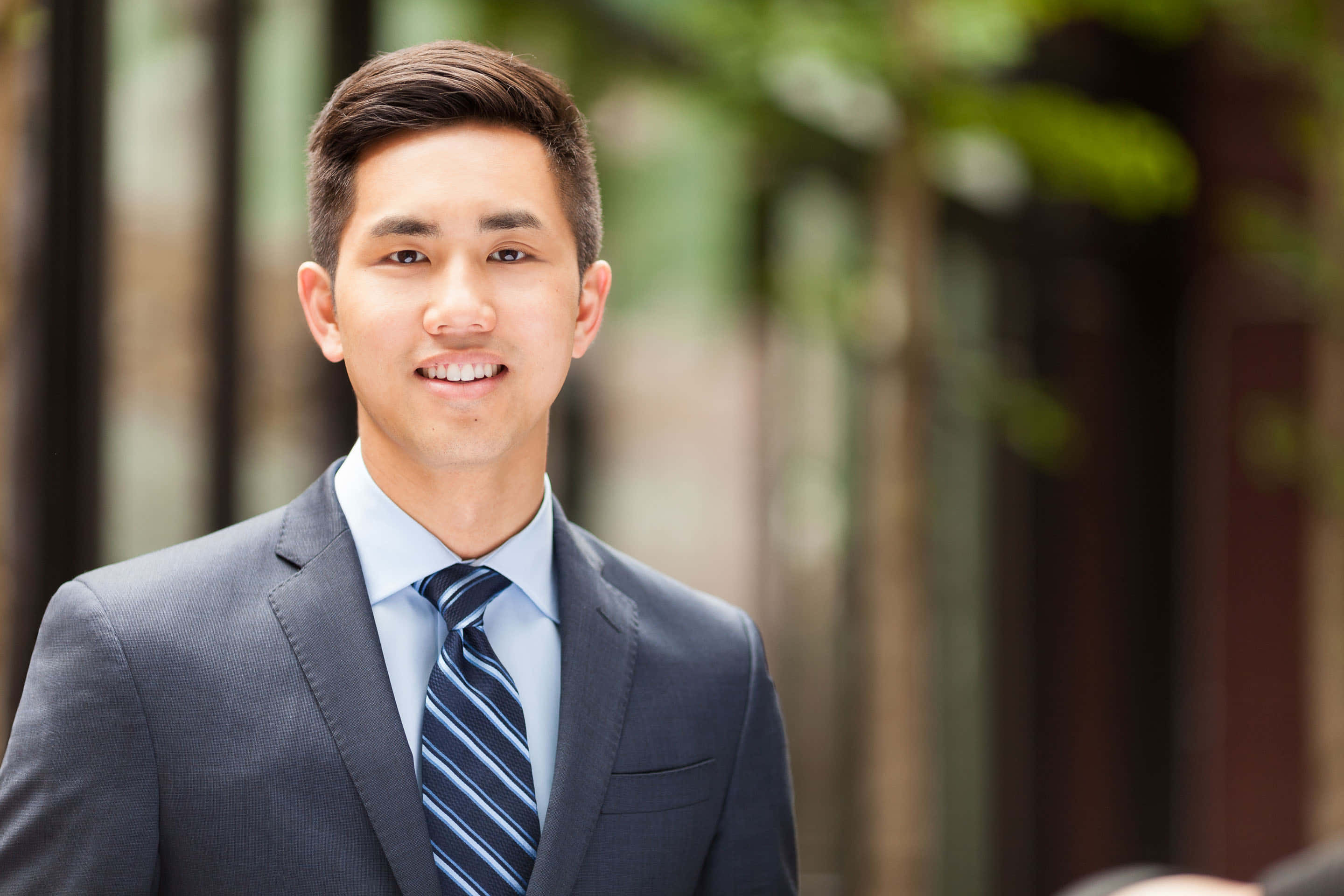 Formal Attire 桌布 壁紙 — 一位微笑的年輕男子身穿俐落灰色西裝和條紋領帶，自信地站在柔和模糊的城市背景前，周圍有綠色植物。