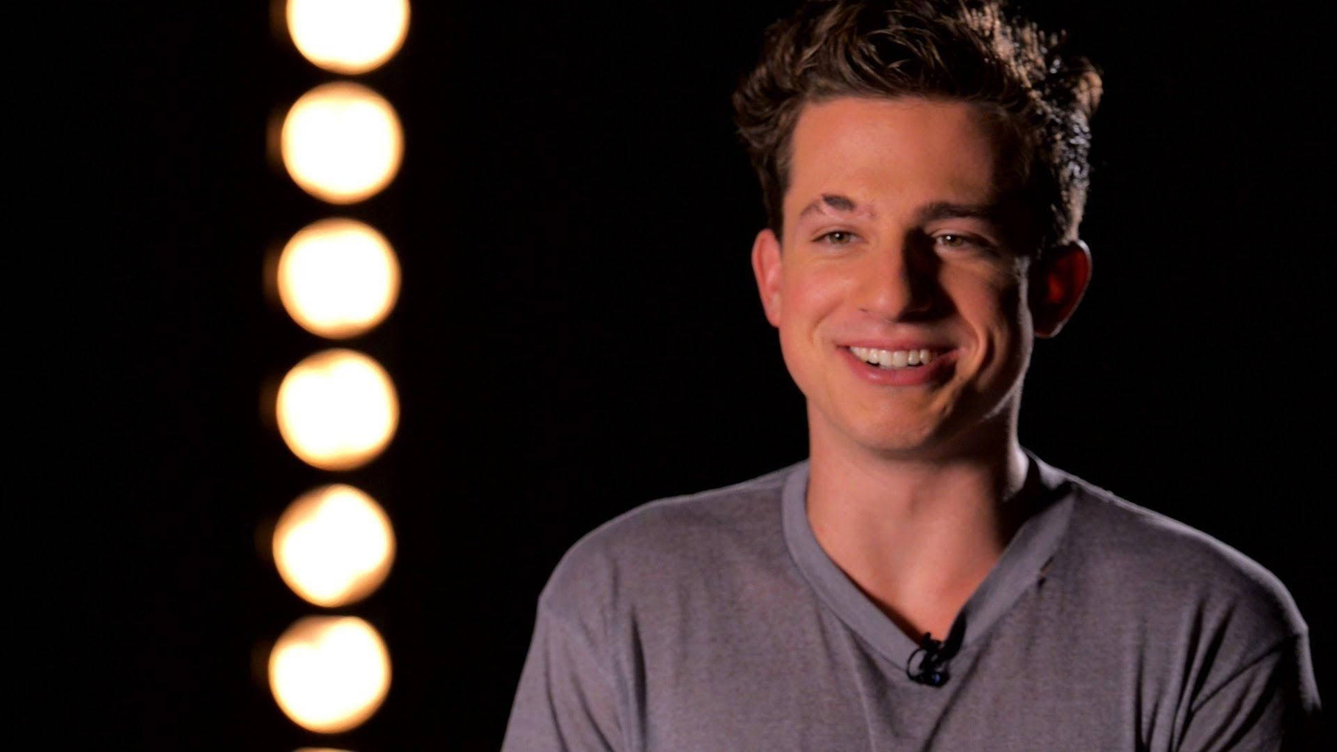 Charlie Puth 桌布 壁紙 — 一位微笑的年輕男子身穿灰色襯衫，站在黑色背景前一排垂直的溫暖發光工作室燈光前。