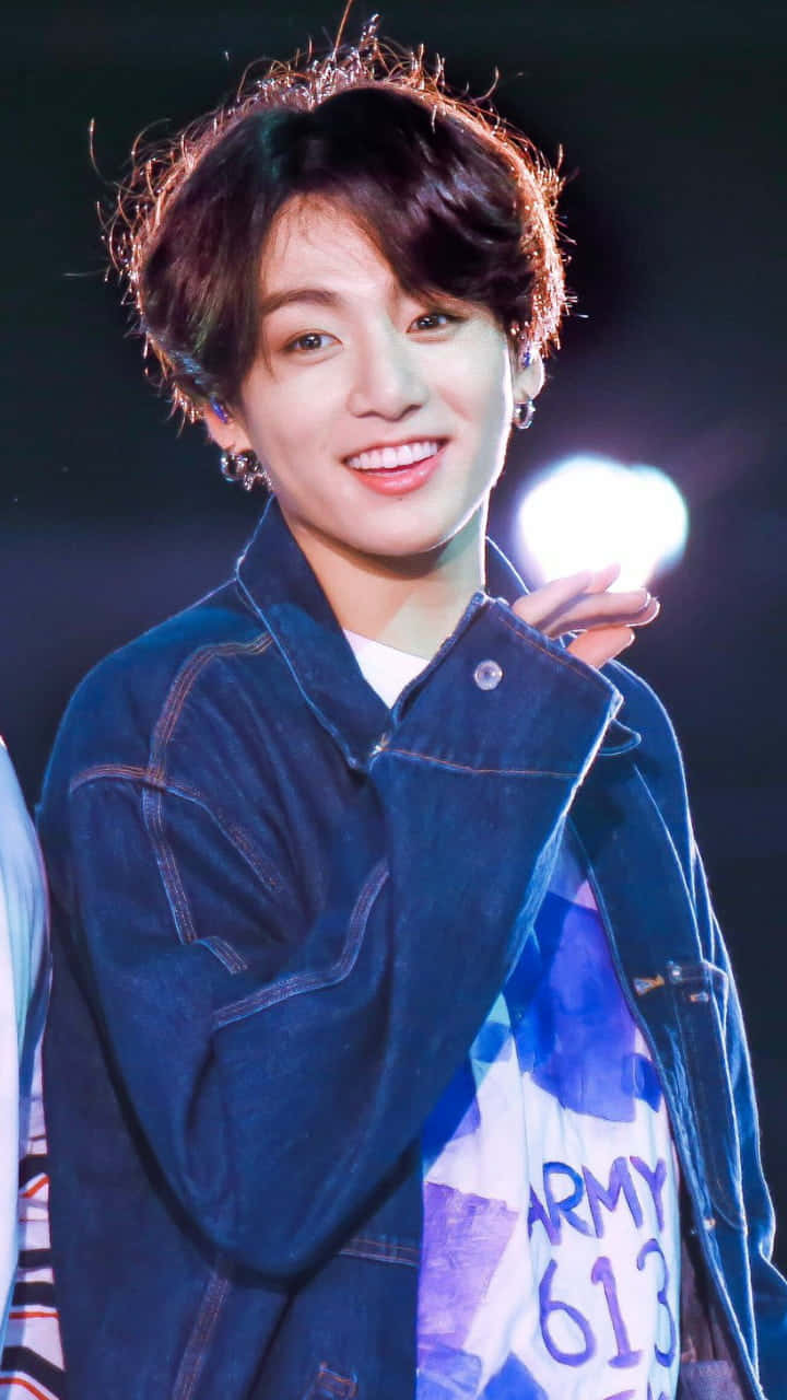 Jungkook 桌布 壁紙 — 一位微笑的年輕男子身穿牛仔外套，在舞台燈光下手持發光球體。