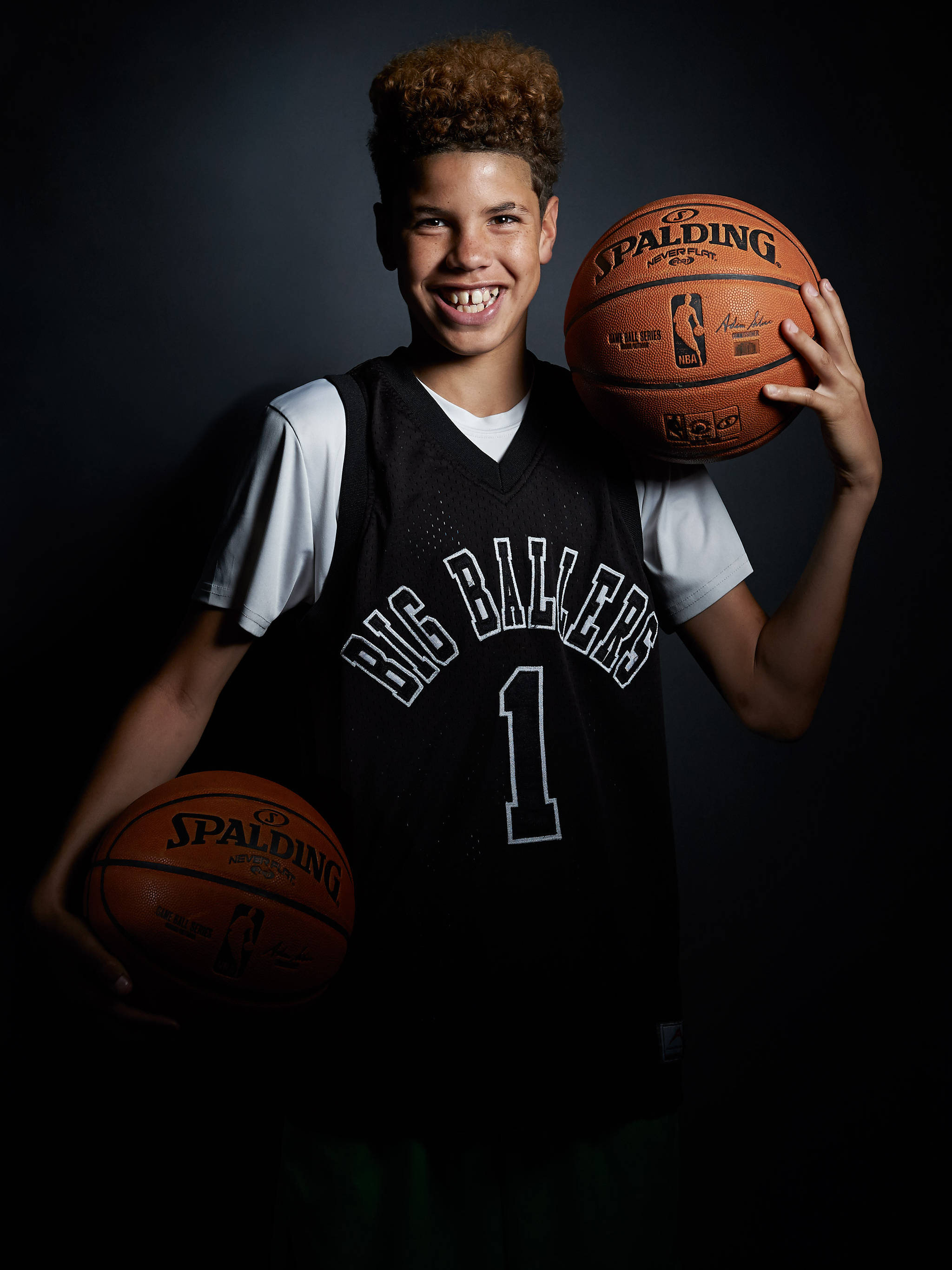 ball 桌布 壁紙 — 一位微笑的年輕籃球運動員身穿黑色 'Big Ballers' 球衣，在深色攝影棚背景下手持兩個 Spalding 籃球。