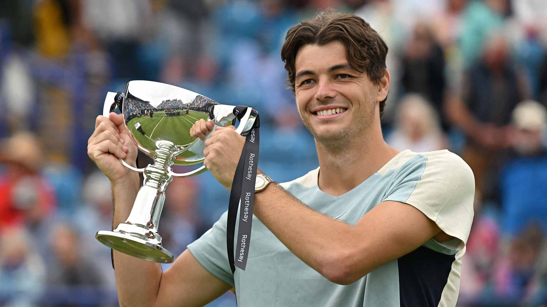 Champion 桌布 壁紙 — 一位微笑的年輕網球運動員自豪地高舉閃亮的銀色獎盃，背景是人群。