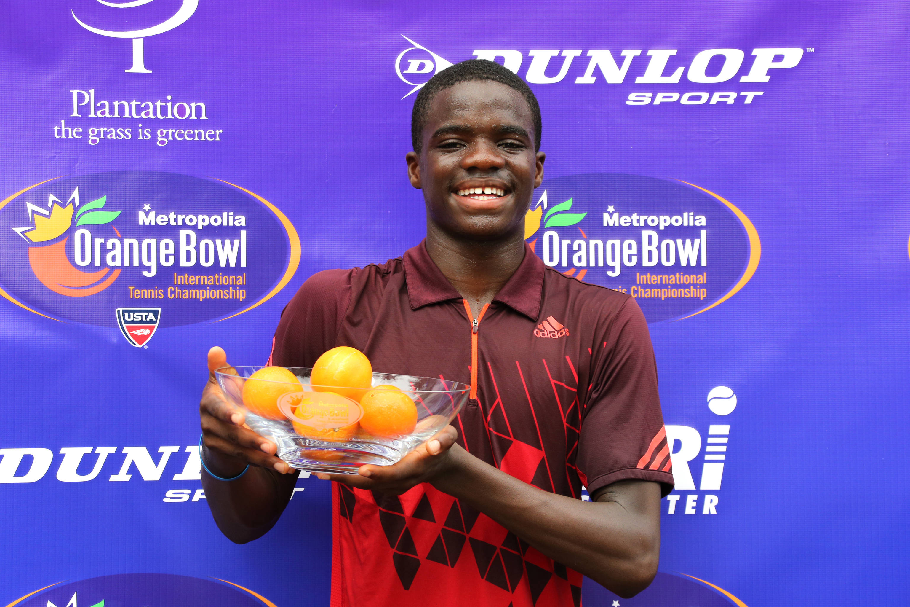 Tennis 桌布 壁紙 — 一位微笑的年輕網球選手自豪地拿著一個裝有柳橙的玻璃碗，背景是紫色的比賽場地。