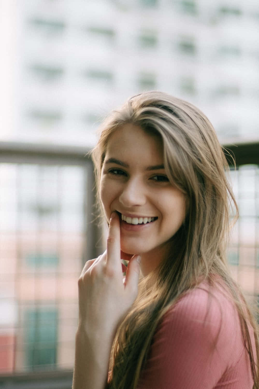 Smile 桌布 壁紙 — 一位微笑的年輕金髮長髮女性，穿著粉紅色上衣，在模糊的城市背景下俏皮地觸碰嘴唇。