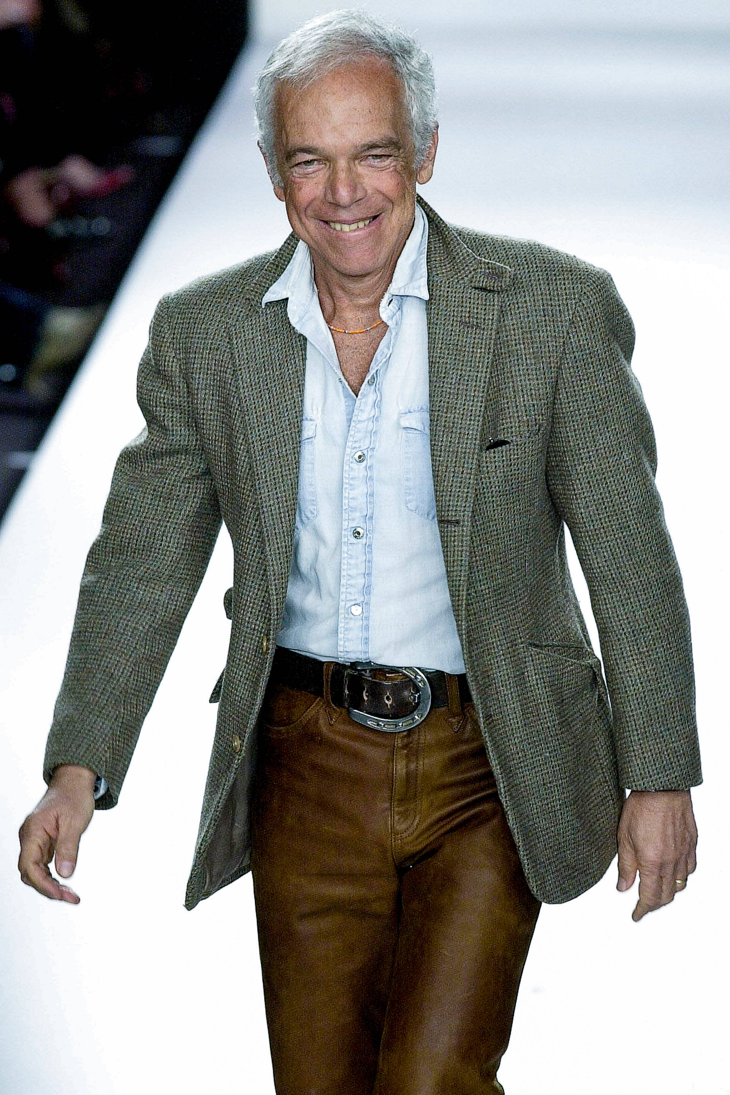 ralph lauren 桌布 壁紙 — 一位微笑的年長男士自信地走在明亮的白色伸展台上，身穿綠色格紋西裝外套和棕色皮褲。