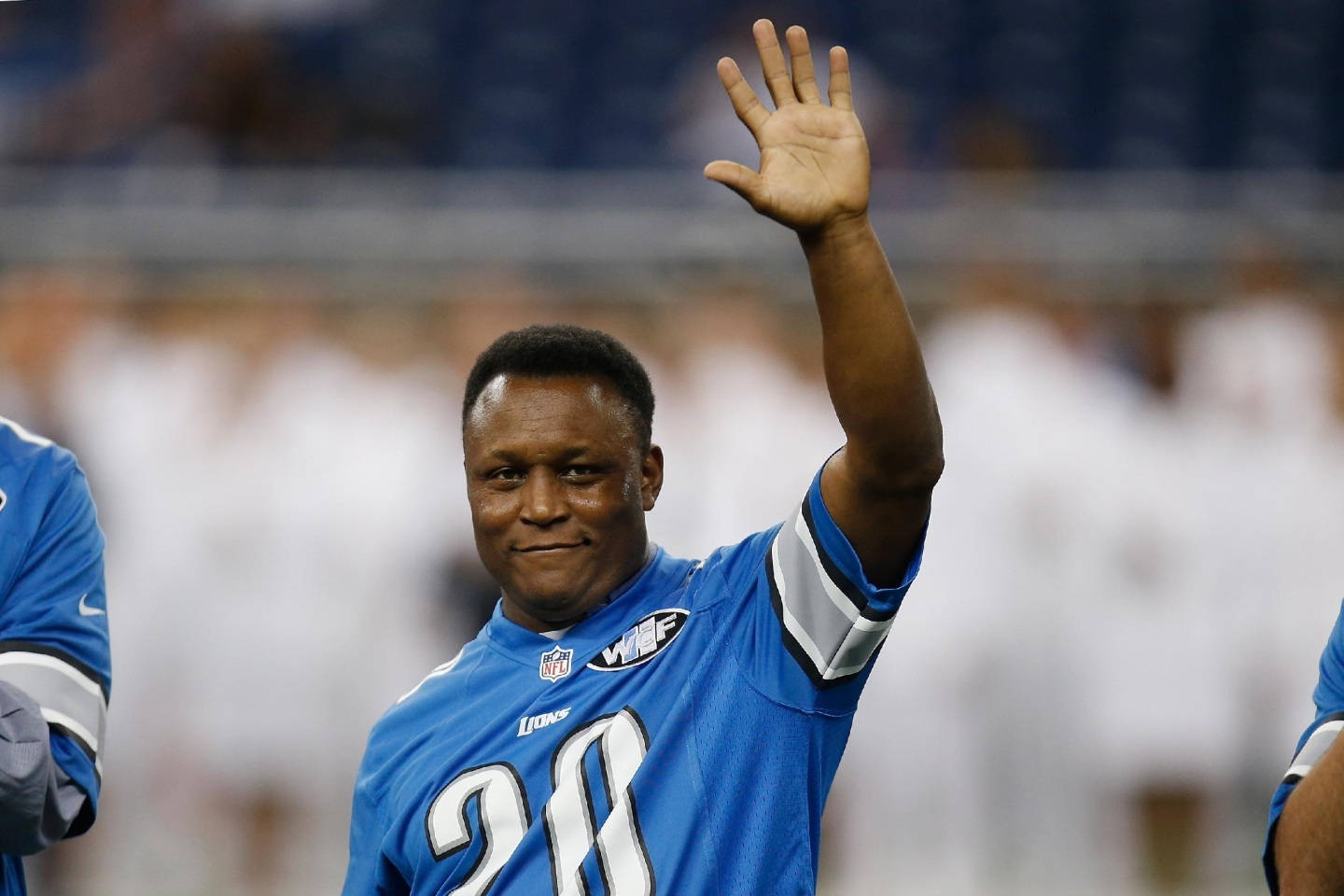 Barry Sanders 桌布 壁紙 — 一位微笑的底特律雄獅美式足球員身穿藍色28號球衣，在明亮燈光下的體育場向球迷揮手。