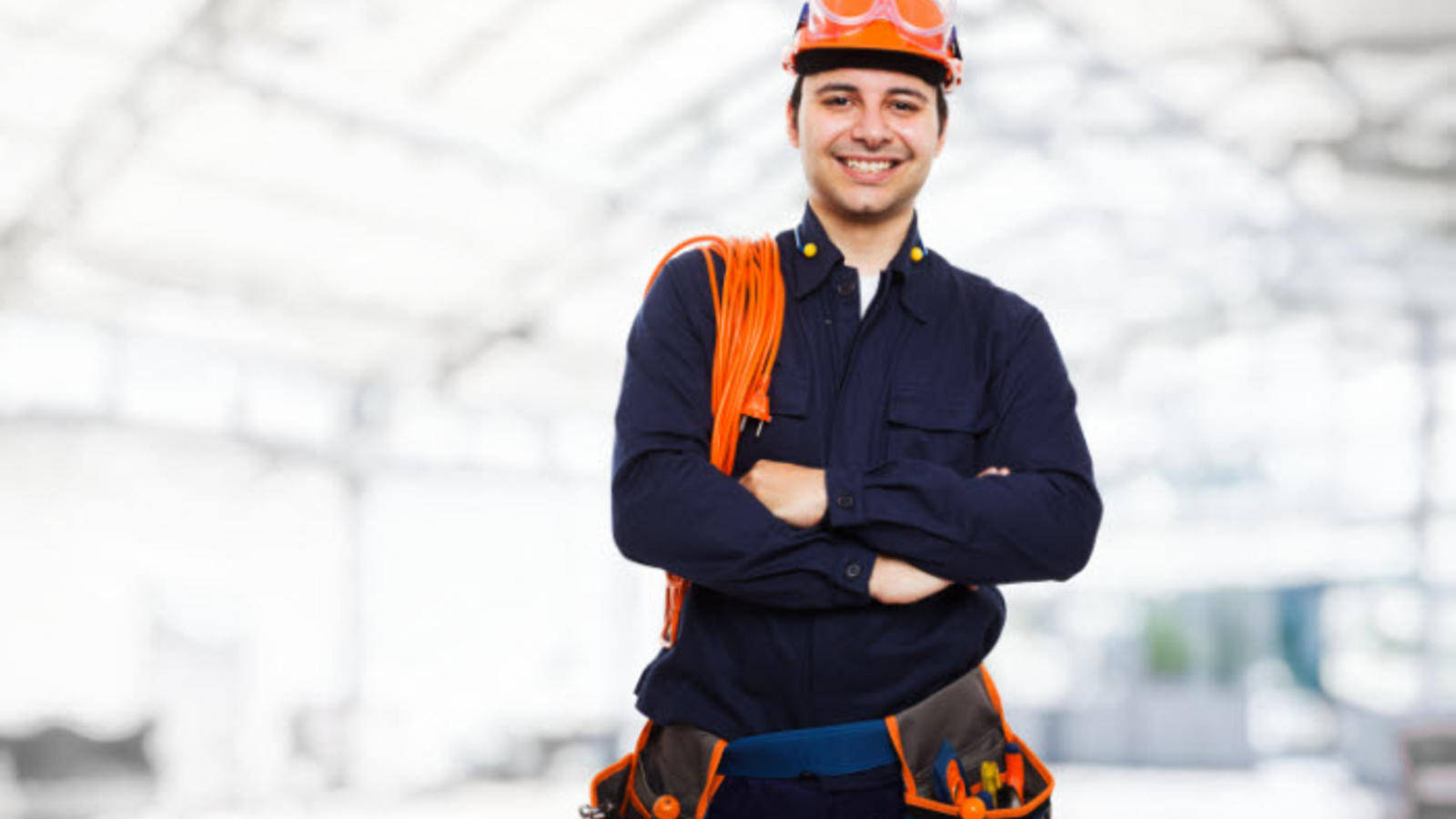 professional 桌布 壁紙 — 一位微笑的建築工人，戴著橙色安全帽和工具腰帶，自信地交叉雙臂站在明亮的工業背景前。