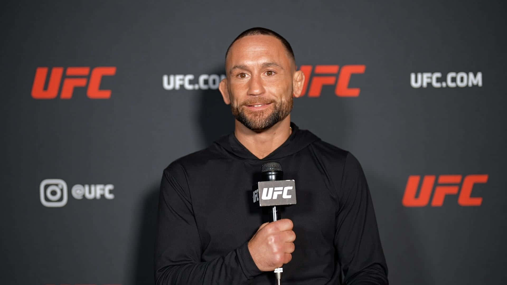UFC 桌布 壁紙 — 一位微笑的戰士穿著黑色連帽衫，在帶有紅色標誌的深灰色背景前握著UFC麥克風。