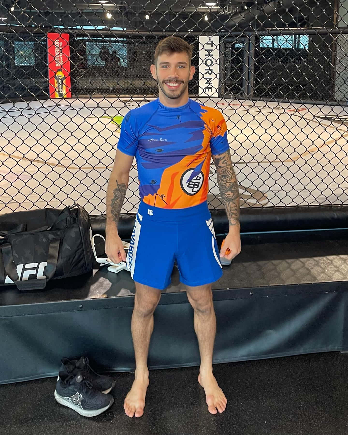 UFC 桌布 壁紙 — 一位微笑的戰士身穿藍色和橙色裝備，赤腳站在MMA籠內，裝備在附近。